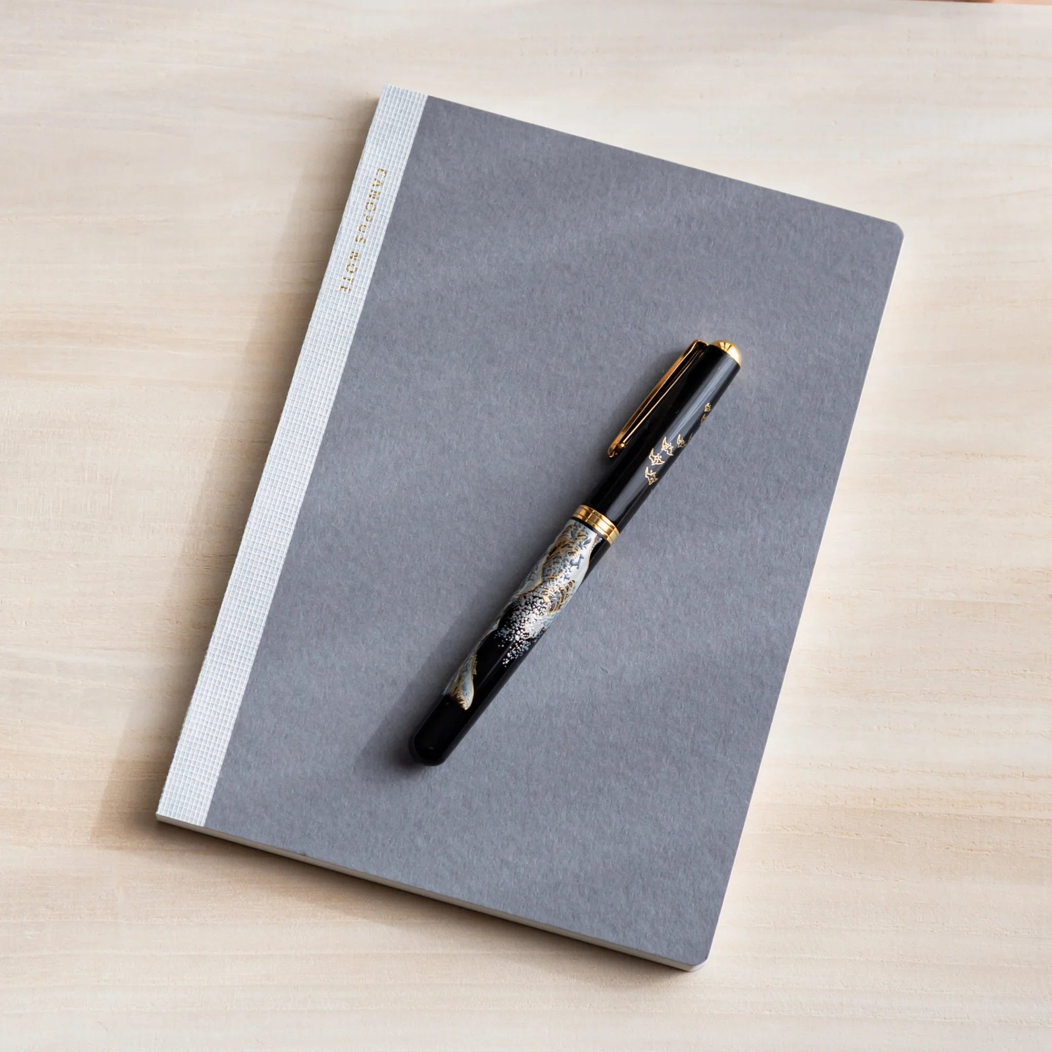 Grey Japanese Notebook with black fountain pen featuring gold floral design on a light wooden desk.