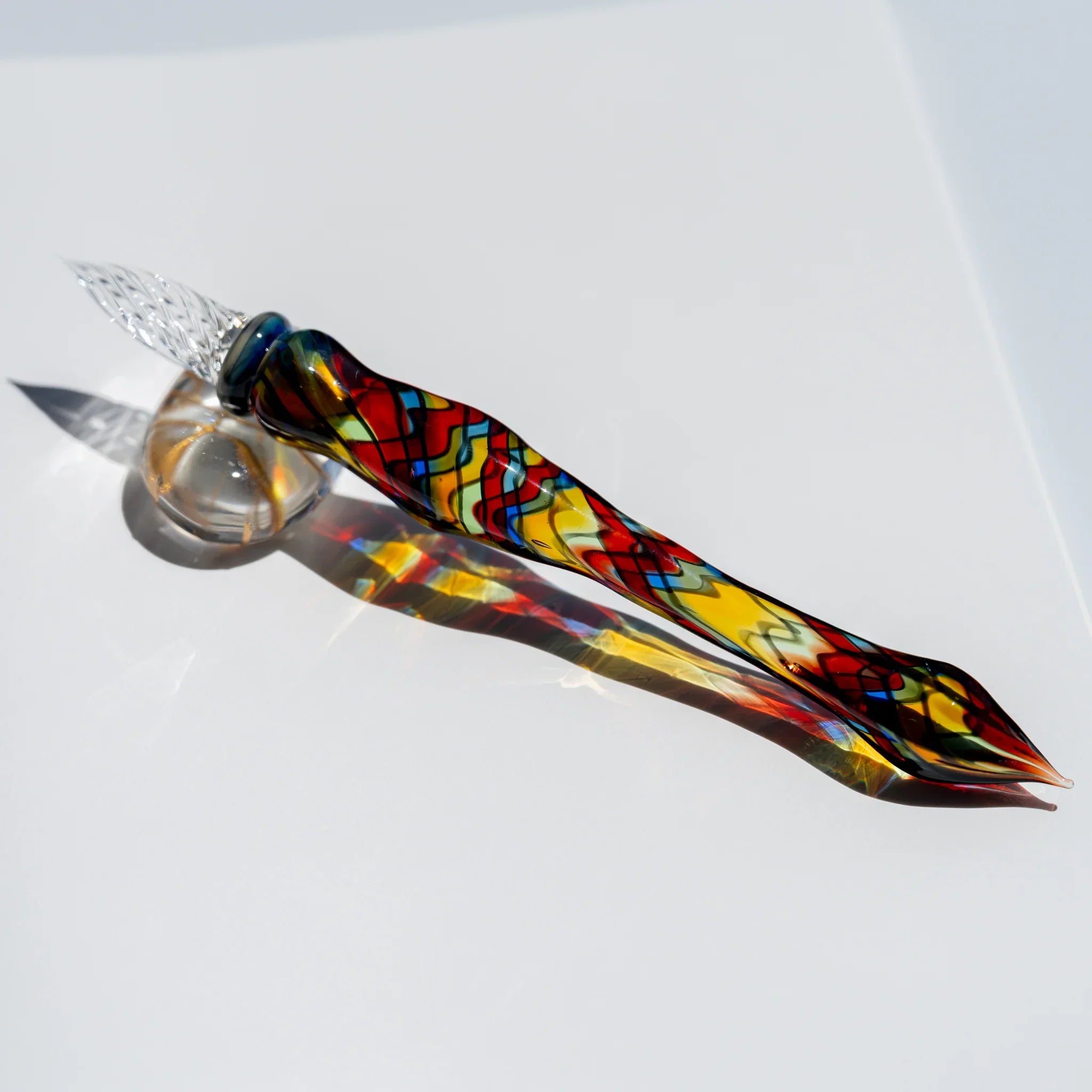 Glass dip pen with a grid-like stained-glass pattern in red, yellow and blue, clear nib on a glass rest