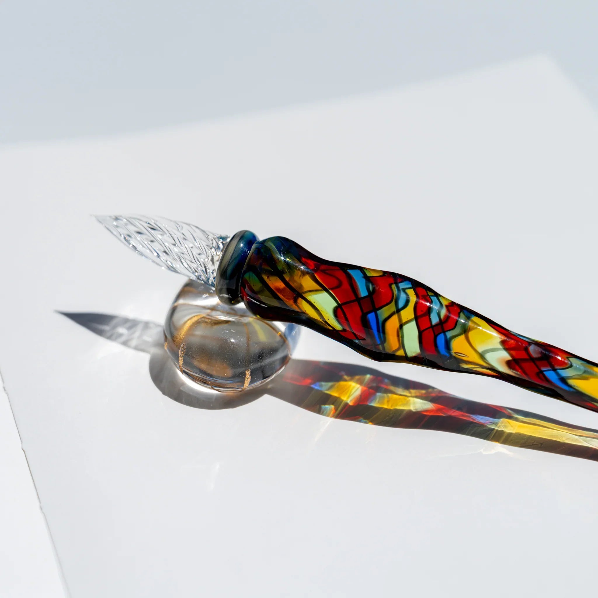 Glass dip pen resting nib-up on a clear glass ball, mosaic body in red, yellow, blue and green casting a colored shadow
