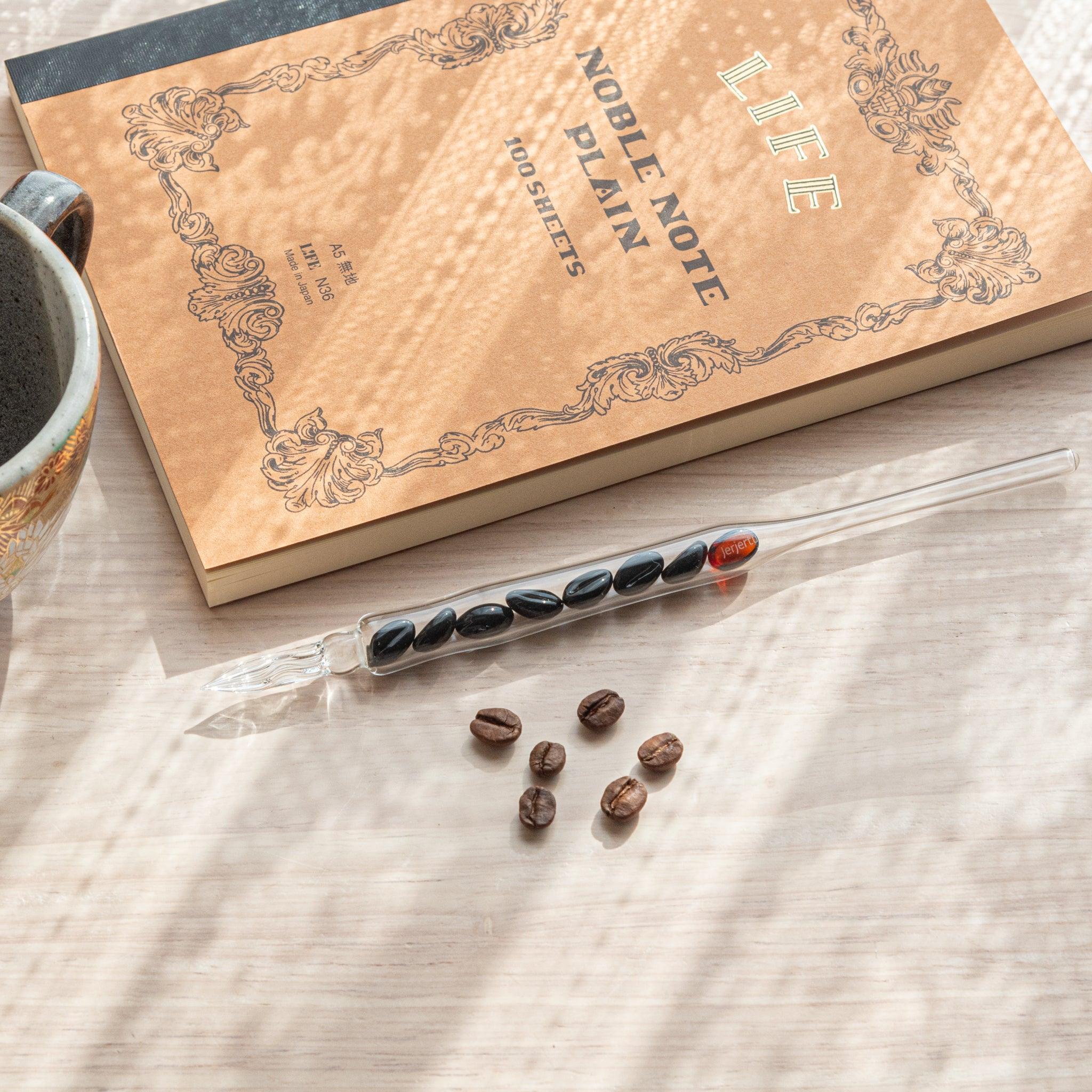 A glass dip pen with coffee bean-shaped glass pieces inside its shaft rests on a wooden surface near coffee beans and a plain notebook.