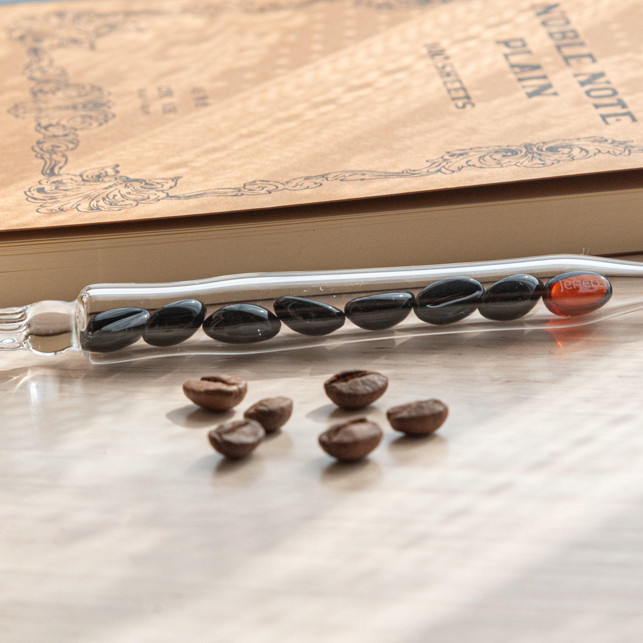 A glass dip pen with coffee bean-shaped glass pieces inside its shaft rests on a wooden surface near coffee beans and a plain notebook.