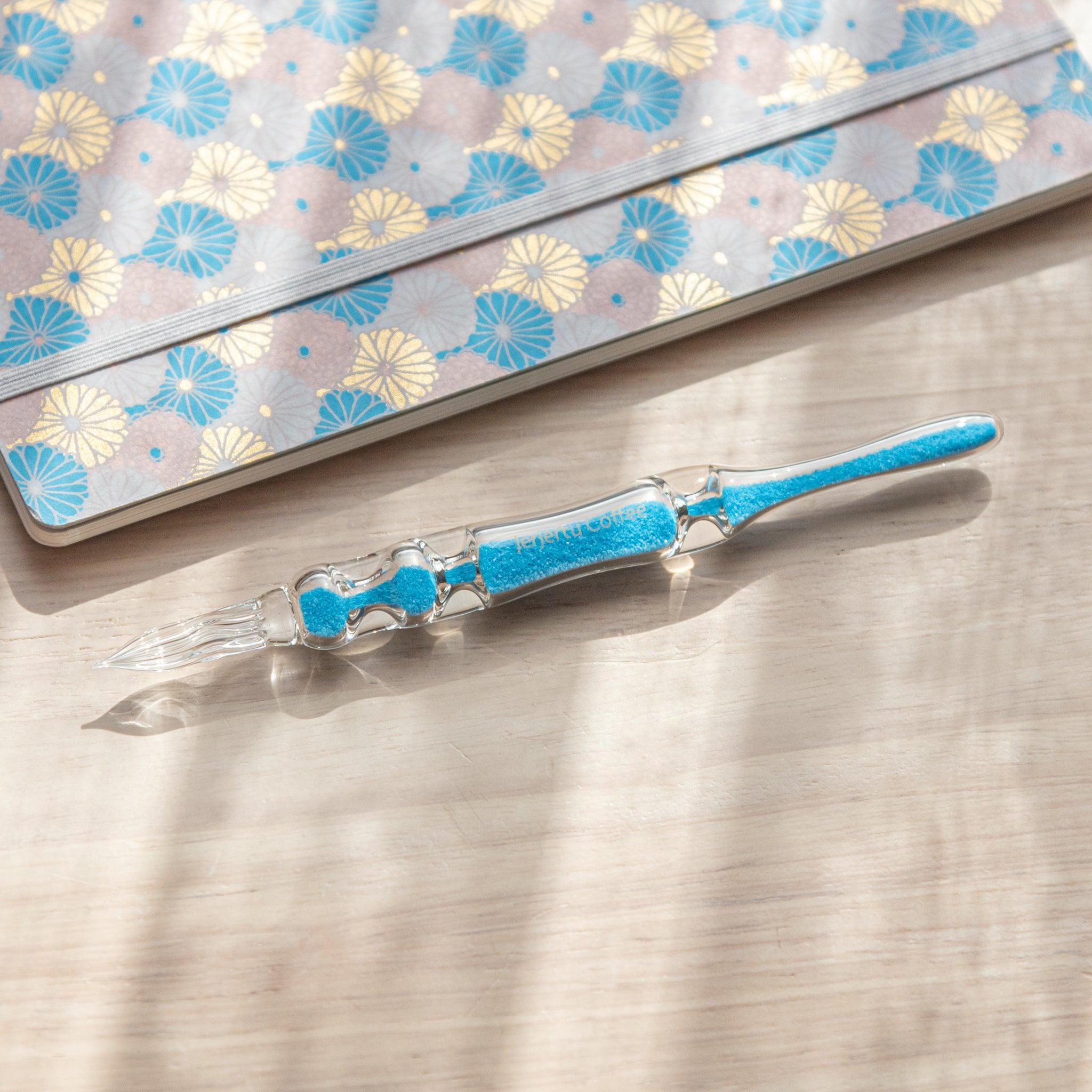 A glass dip pen with blue sand-like particles inside its shaft, engraved with "Jerjertu coffee," rests on a wooden surface near a pastel floral-patterned notebook. A bottle of blue ink is partially visible on the left.