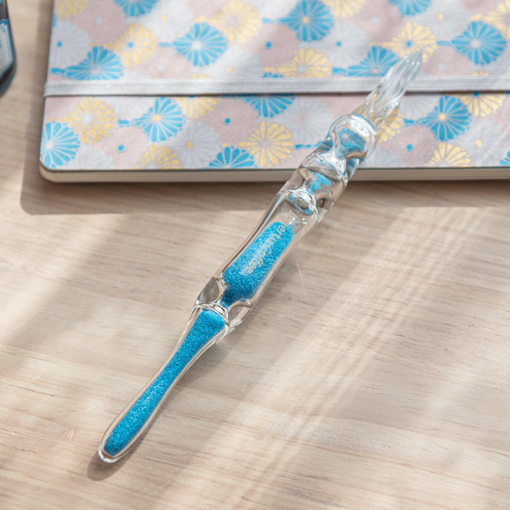 A glass dip pen with blue sand-like particles inside its shaft, engraved with "Jerjertu coffee," rests on a wooden surface near a pastel floral-patterned notebook. A bottle of blue ink is partially visible on the left.