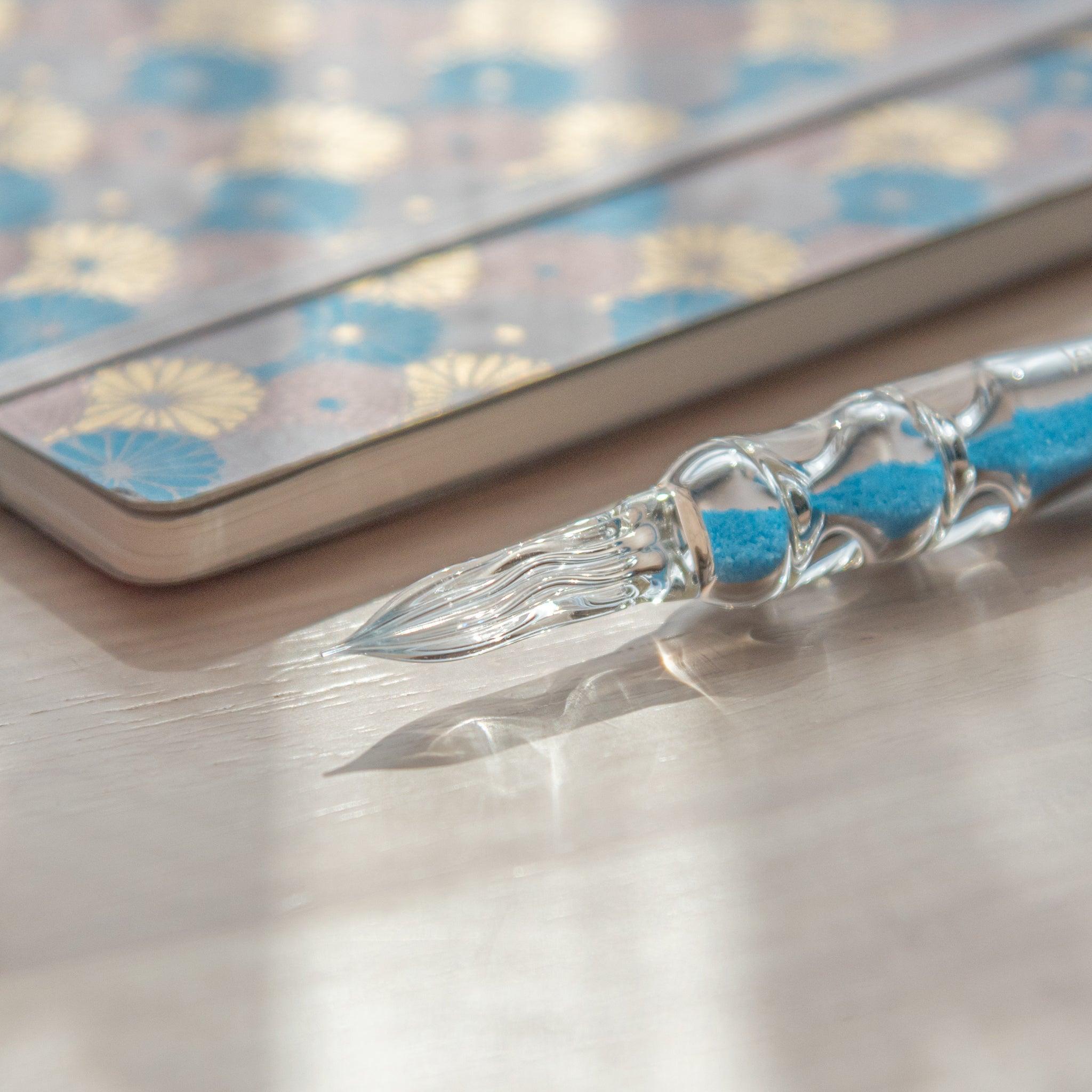 A glass dip pen with blue sand-like particles inside its shaft, engraved with "Jerjertu coffee," rests on a wooden surface near a pastel floral-patterned notebook. A bottle of blue ink is partially visible on the left.