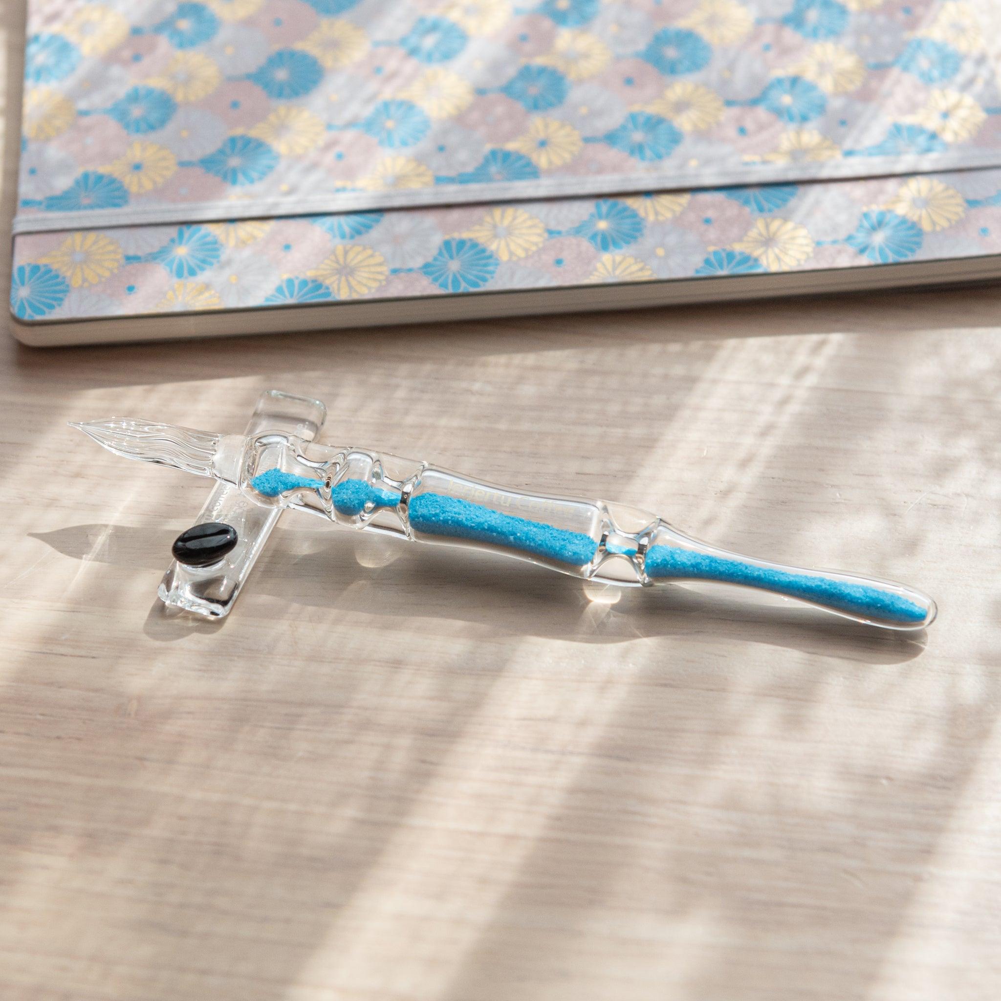 A glass dip pen with blue sand-like particles inside its shaft, engraved with "Jerjertu coffee," rests on a wooden surface near a pastel floral-patterned notebook. A bottle of blue ink is partially visible on the left.