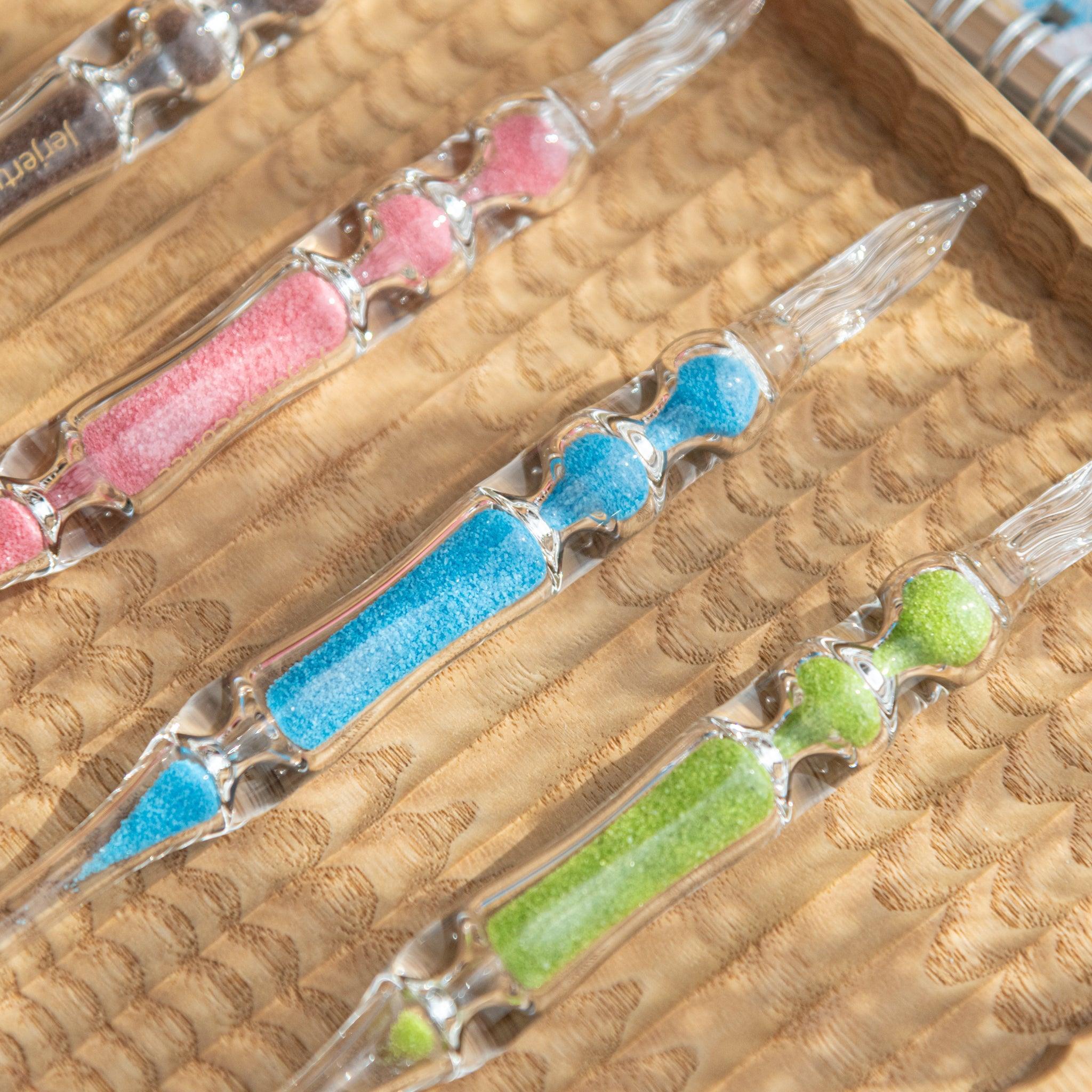 A close-up view of four glass dip pens with sand-like particles inside their shafts in pink, blue, green, and brown. The pens are arranged neatly on a carved wooden tray, highlighting the vibrant colors and transparent glass details.