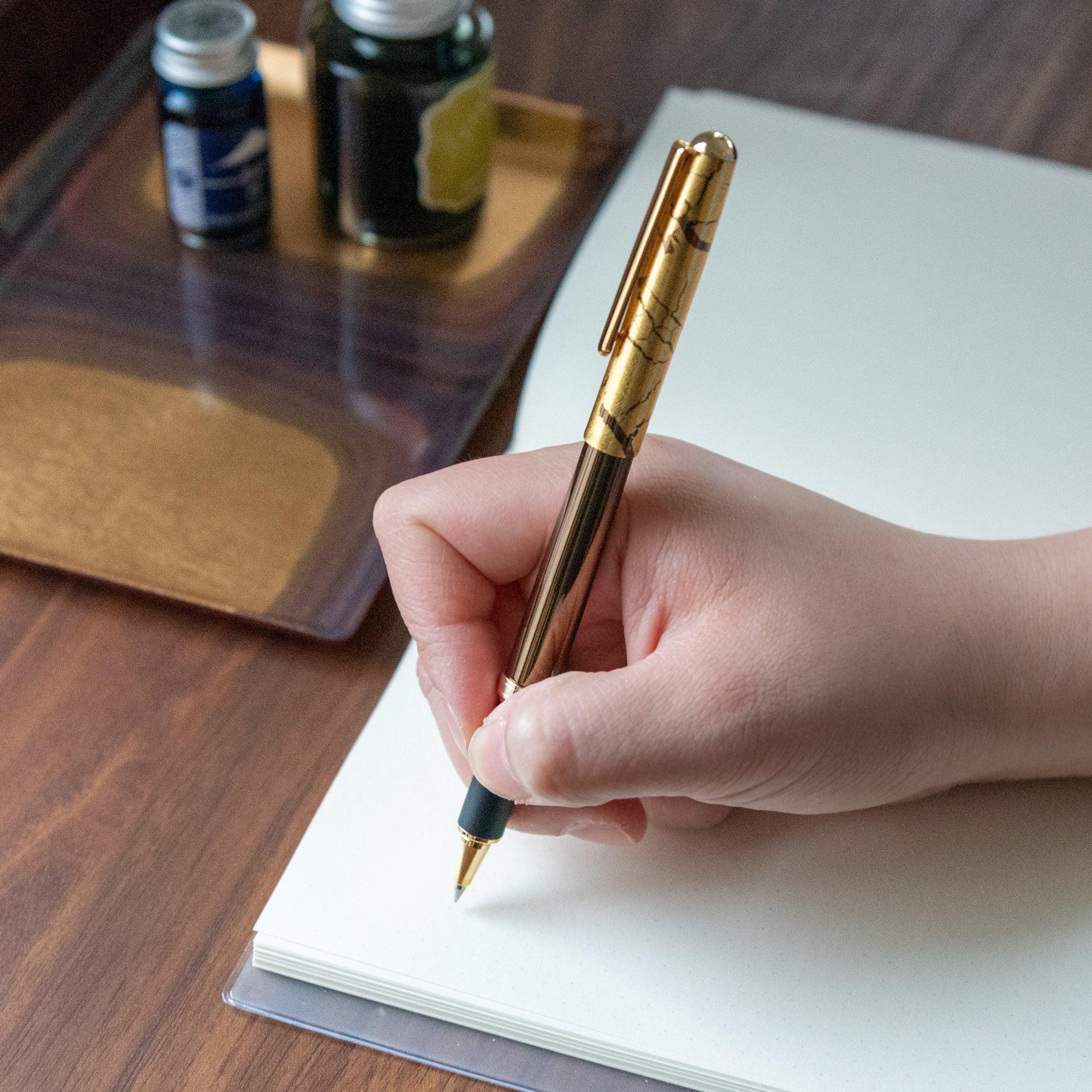 A person writing with a brown pen featuring a gold crack pattern cap, with ink bottles and a wooden tray in the background.