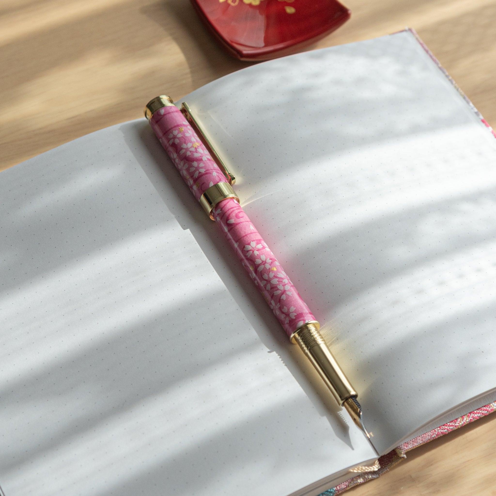 Pink sakura-themed fountain pen with gold accents resting on an open dotted notebook under soft sunlight.