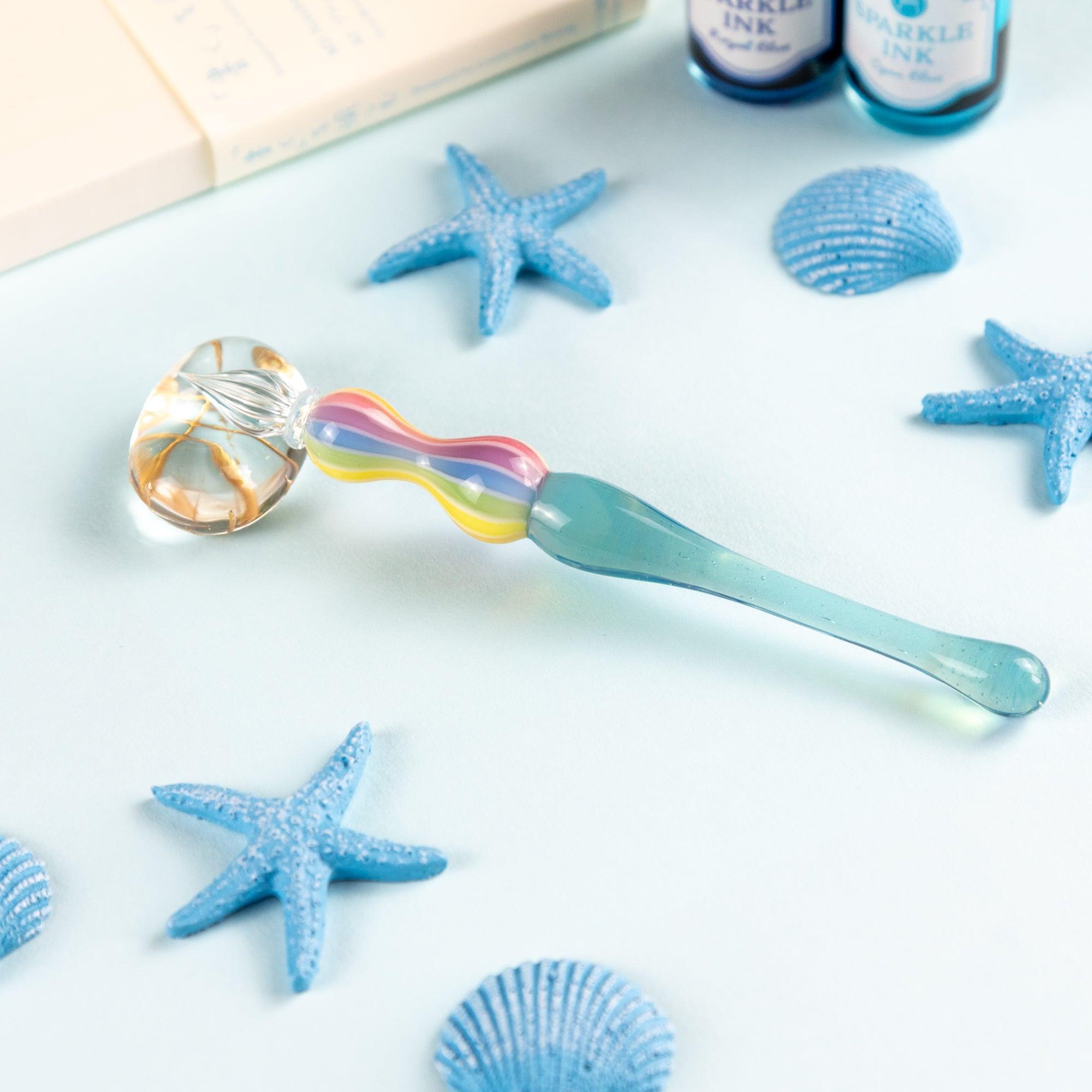 Rainbow-colored glass pen resting on a clear glass pen rest with blue starfish and seashell decor. Perfect for Japanese stationery enthusiasts.