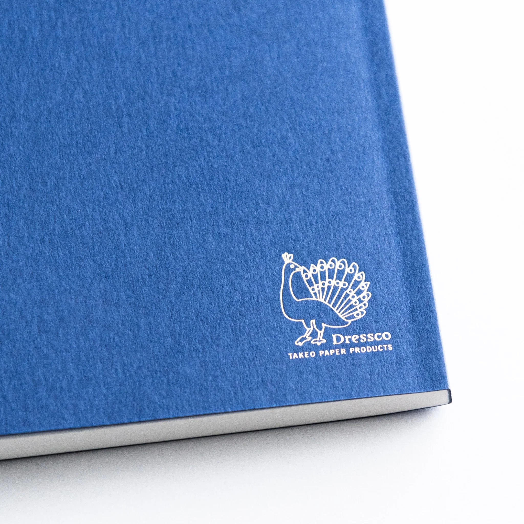 Close-up of white Dressco peacock logo embossed on textured blue cover of Japanese Notebook.
