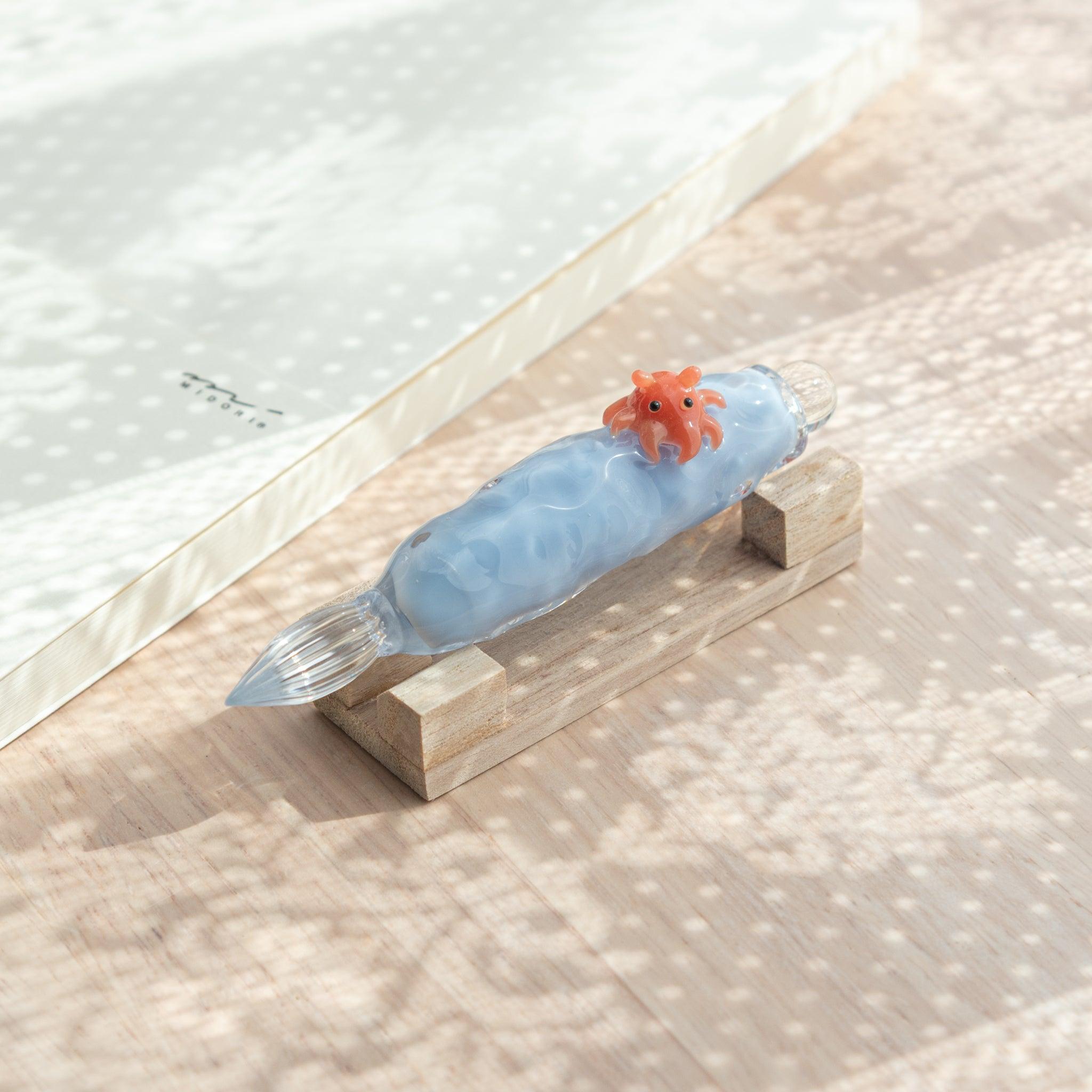 Dumbo Octopus Glass Dip Pen on a wooden holder, glowing in soft light with lace-like shadows on a wooden surface.