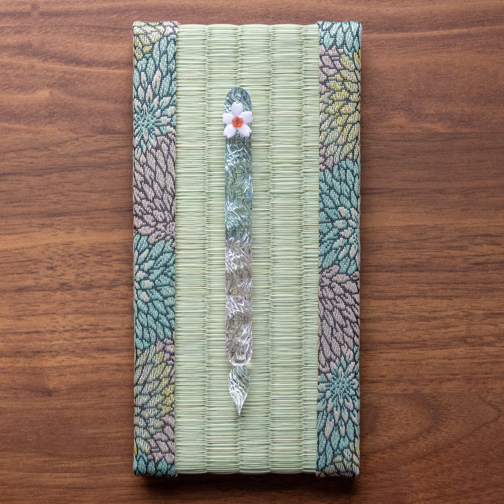 Glass pen with a sakura flower design placed on a tatami pen tray bordered with floral-patterned fabric, showcasing exquisite Japanese stationery.