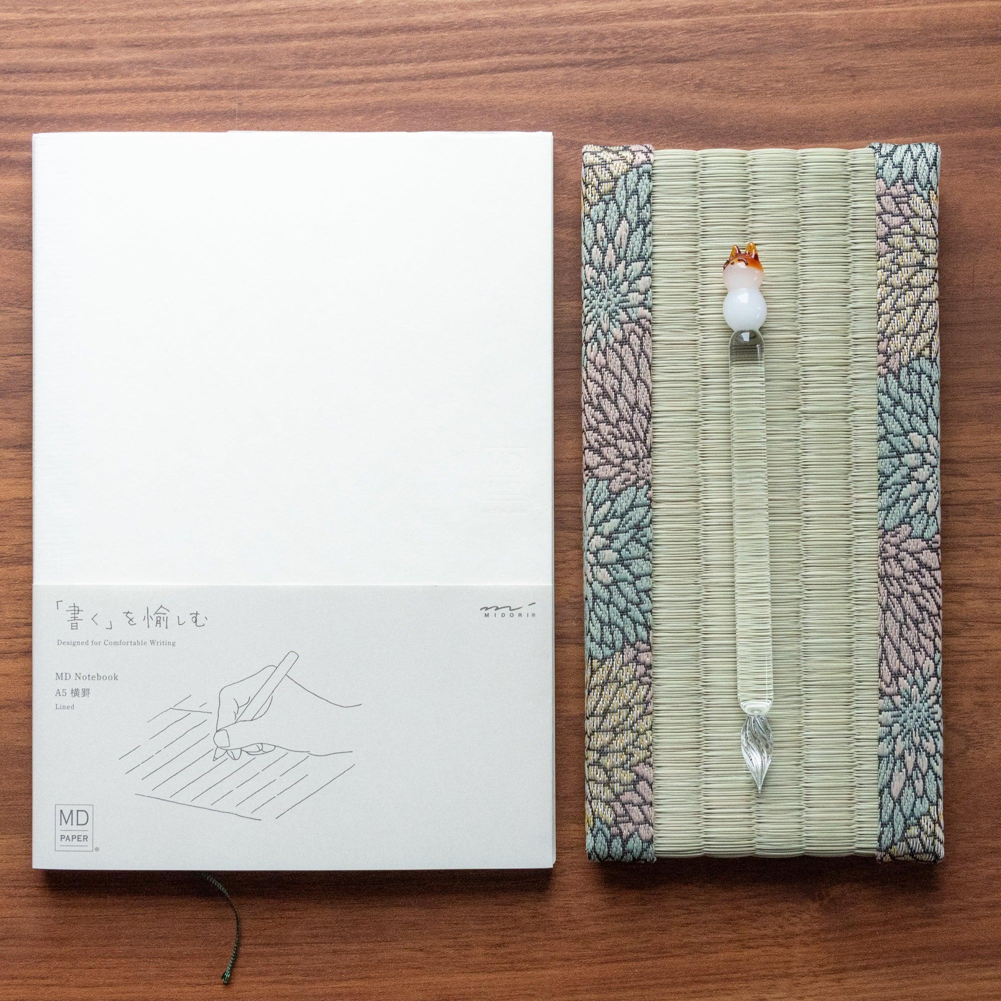 A minimalist MD notebook placed beside a tatami pen tray holding a glass pen with a fox motif, emphasizing the elegance of Japanese stationery.