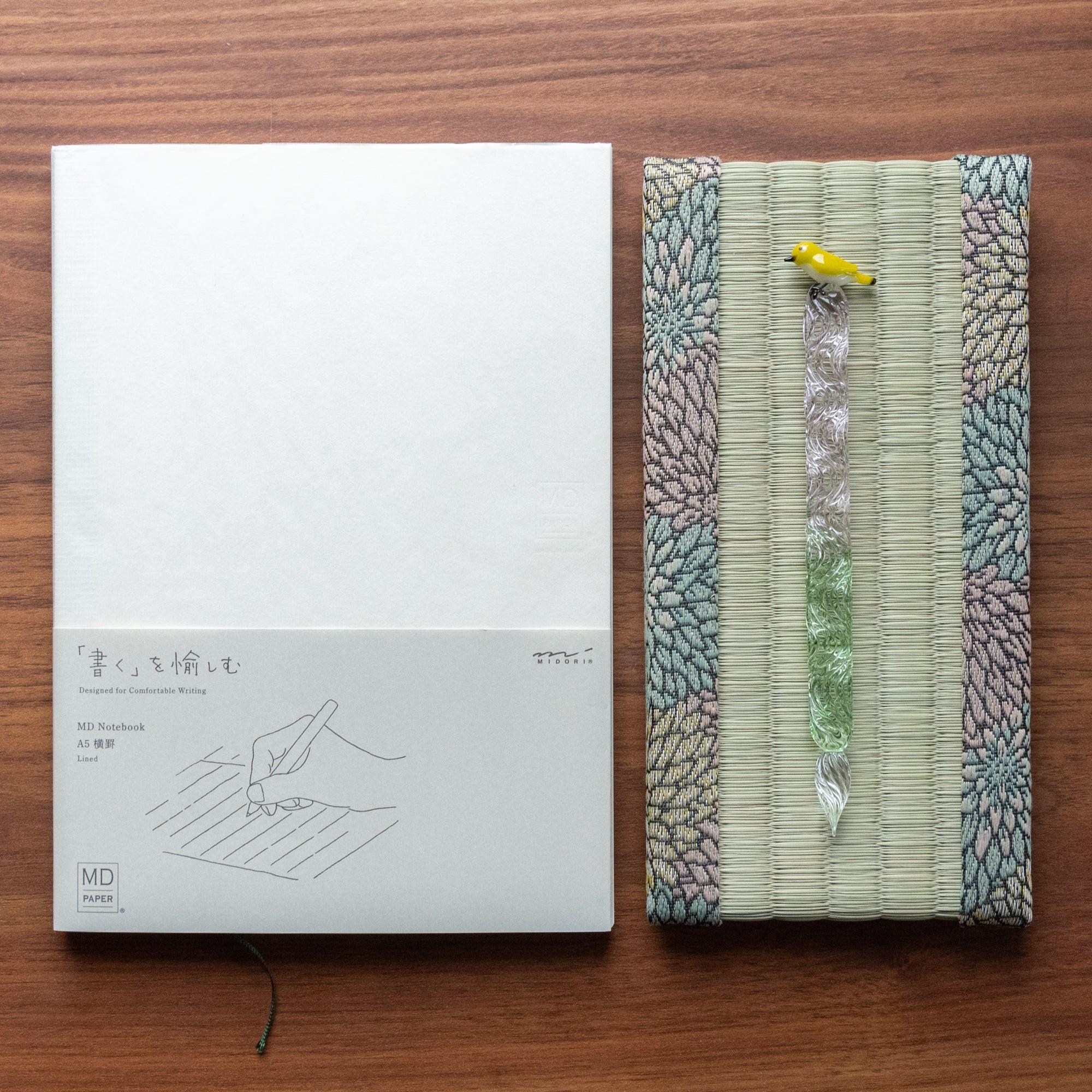 A minimalist MD notebook placed beside a tatami pen tray holding a glass pen with a yellow bird motif, showcasing elegant Japanese stationery.