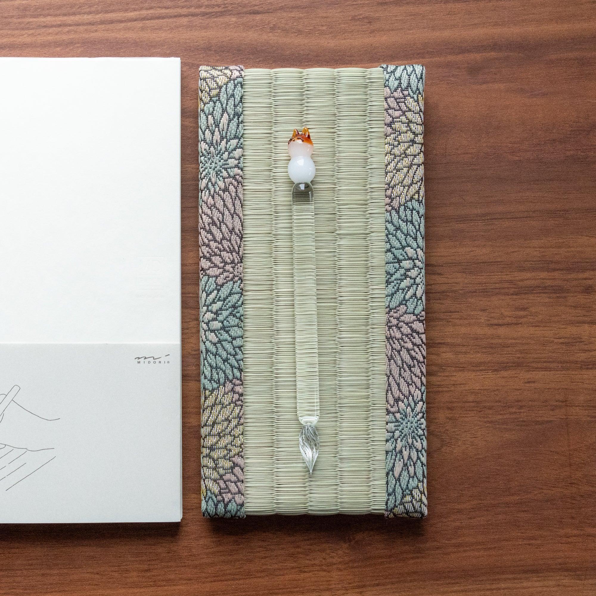 Overhead flat lay of Chrysanthemum Tatami Pen Tray with glass pen placed vertically and Midori notebook beside it