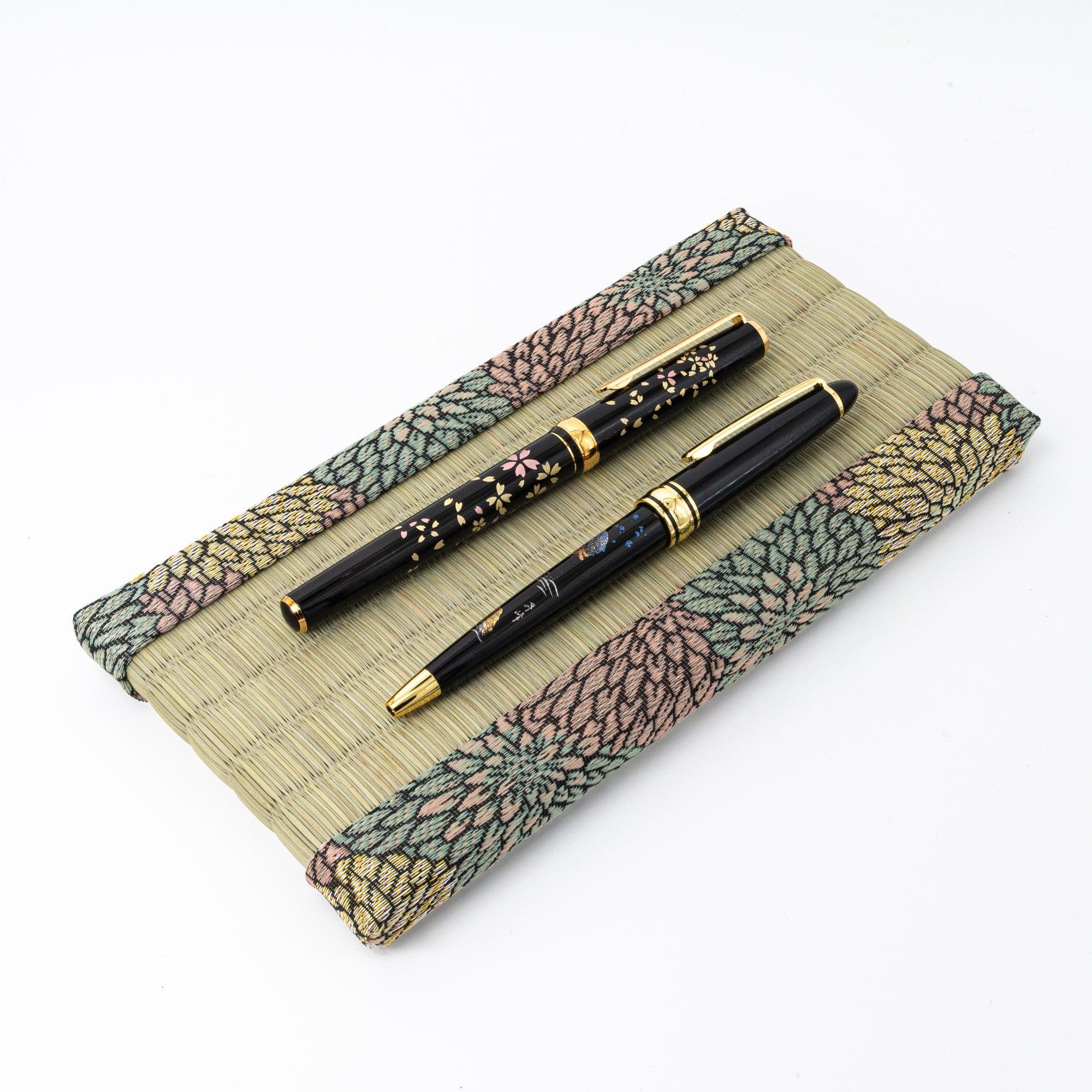Two black lacquer pens with gold floral motifs resting on a tatami pen tray with floral-patterned borders, emphasizing refined Japanese stationery.