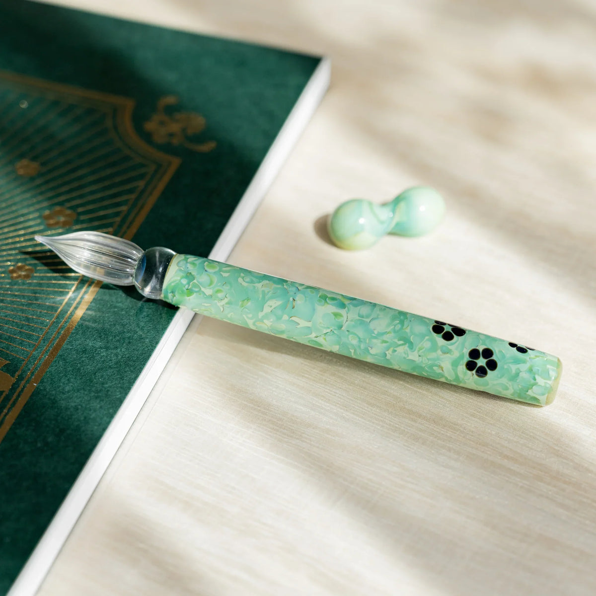 Emerald Flower glass dip pen on a white page with its two-sphere green glass pen rest to one side