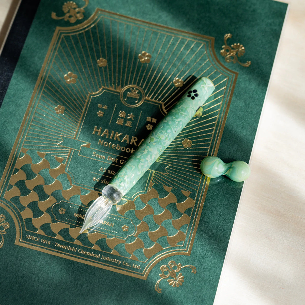 Emerald Flower glass dip pen and pen rest on a green HAIKARA notebook cover with gold foil printing