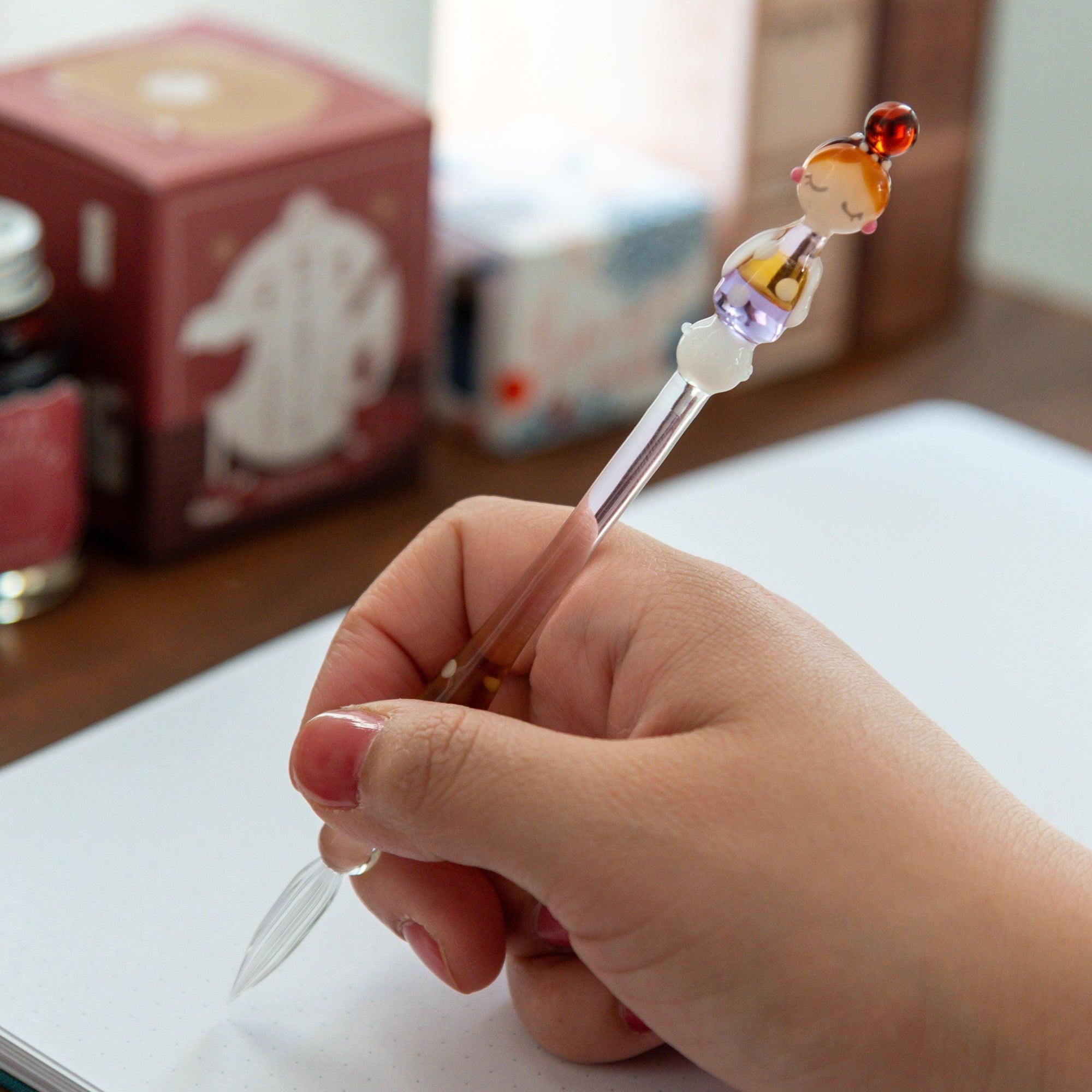 Hand holding the Fashionable Lady Marun-chan glass pen, showing the pen in use on a white notebook with a cozy tea container in the background