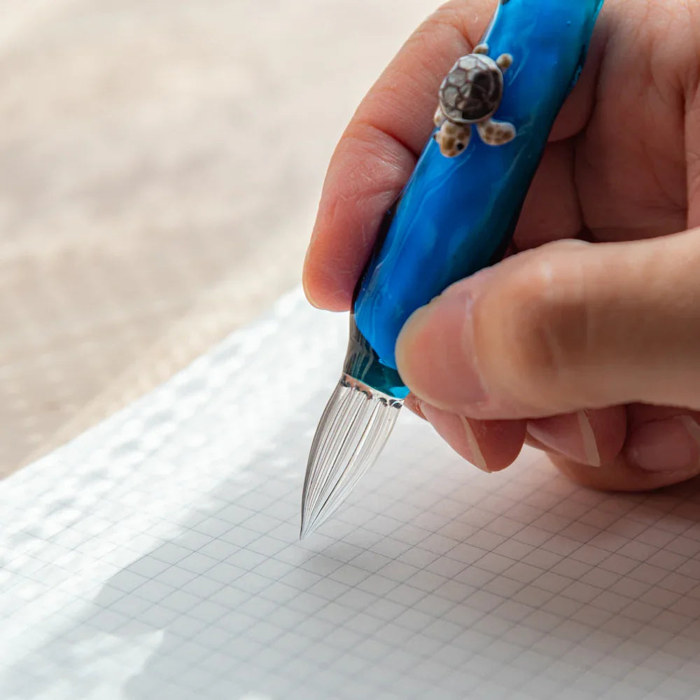 Close-up of a hand writing with a blue glass dip pen featuring a small sea turtle decoration on the barrel.