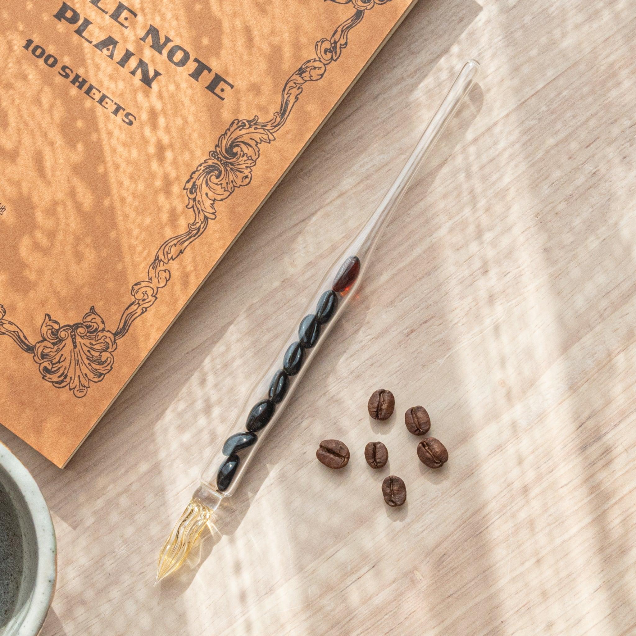A glass dip pen with coffee bean-shaped glass pieces inside its shaft and a coffee-stained colored nib rests on a wooden surface near coffee beans and a plain notebook.