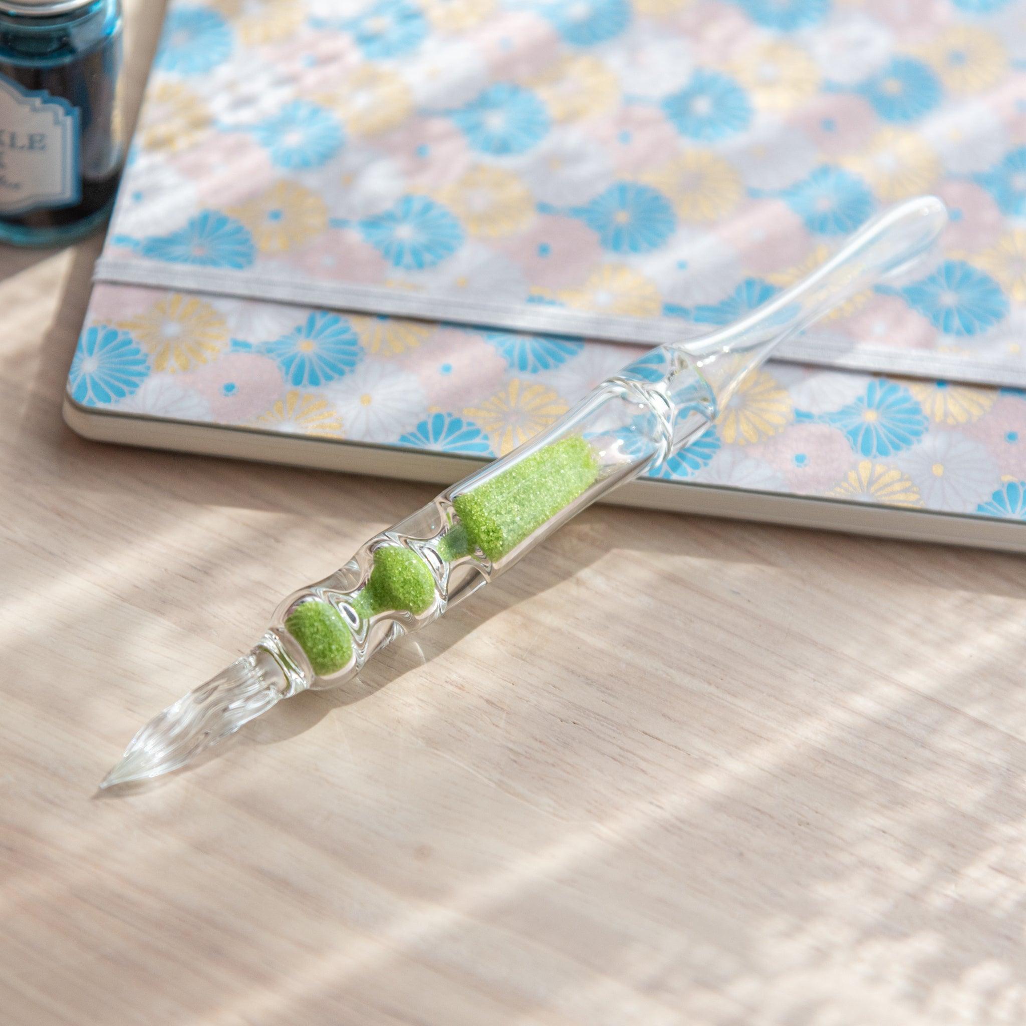 A glass dip pen with green sand-like particles inside its shaft rests on a wooden surface next to a pastel floral-patterned notebook. The pen's transparent design showcases the vibrant green particles inside.