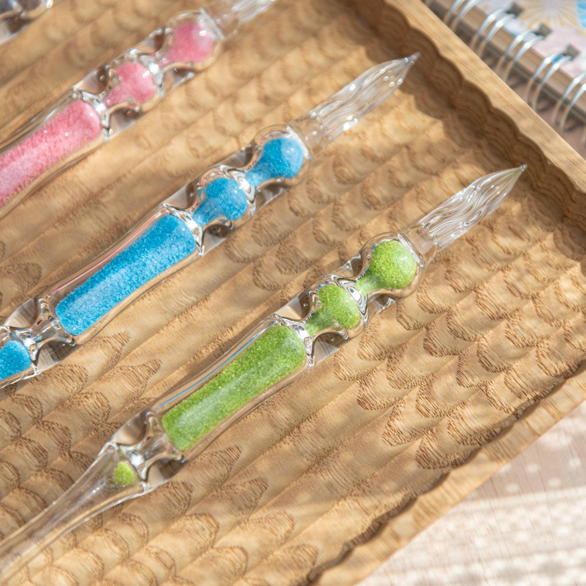 Four glass dip pens with sand-like particles inside their shafts in pink, blue, green, and brown are arranged neatly on a carved wooden tray. The bright colors of the sand-like particles contrast with the clear glass of the pens.