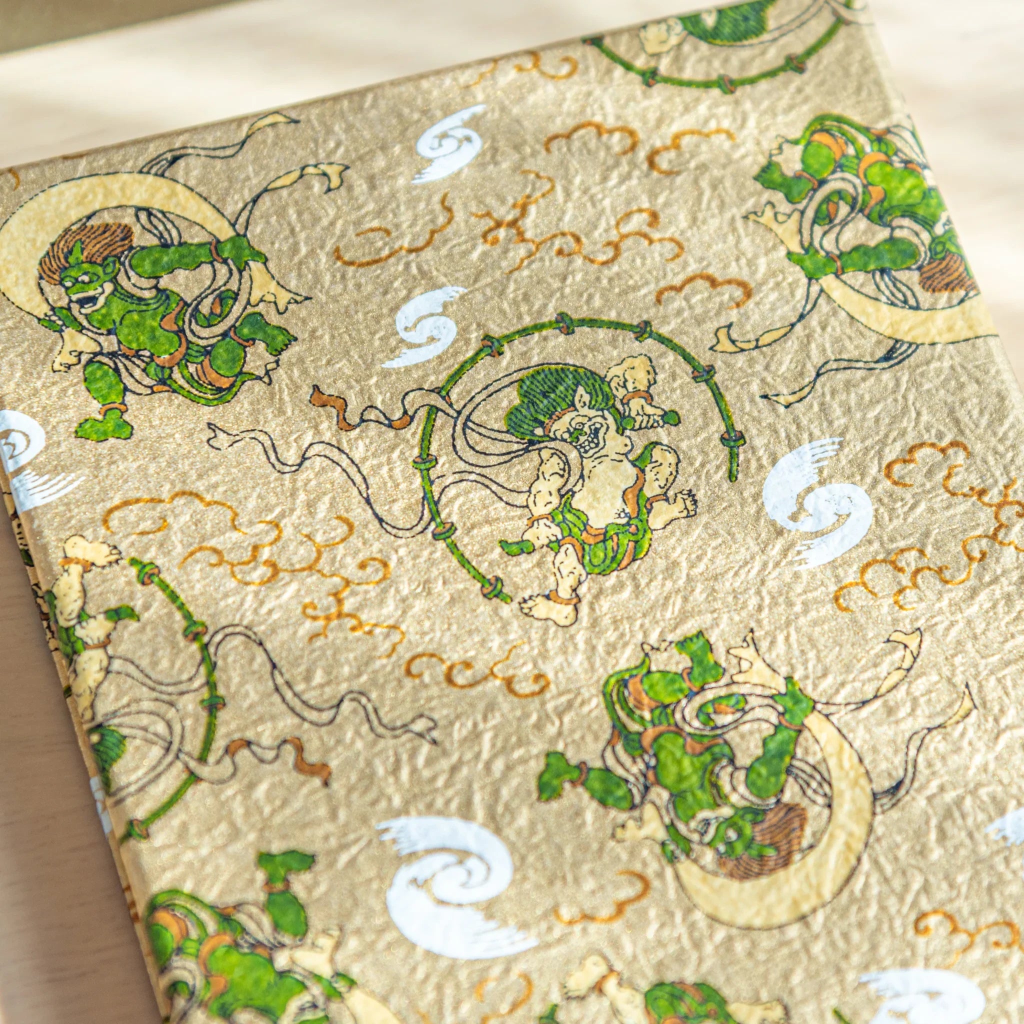 Gold washi goshuincho stamp book cover with Fujin and Raijin figures in green, white swirls and amber clouds, angled close-up