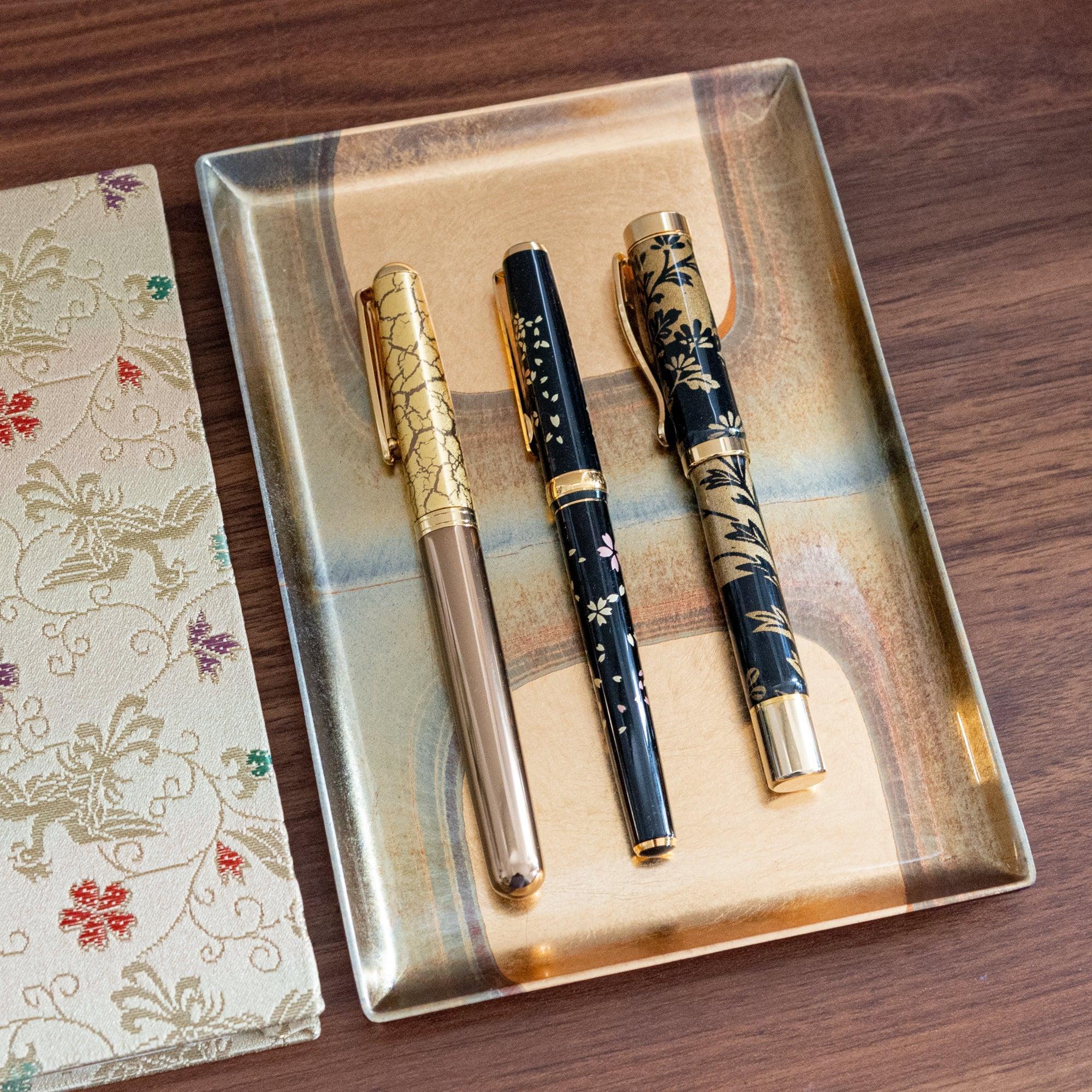 Three Japanese stationery pens with gold accents displayed on a lacquered tray, alongside a fabric notebook cover with a traditional design.