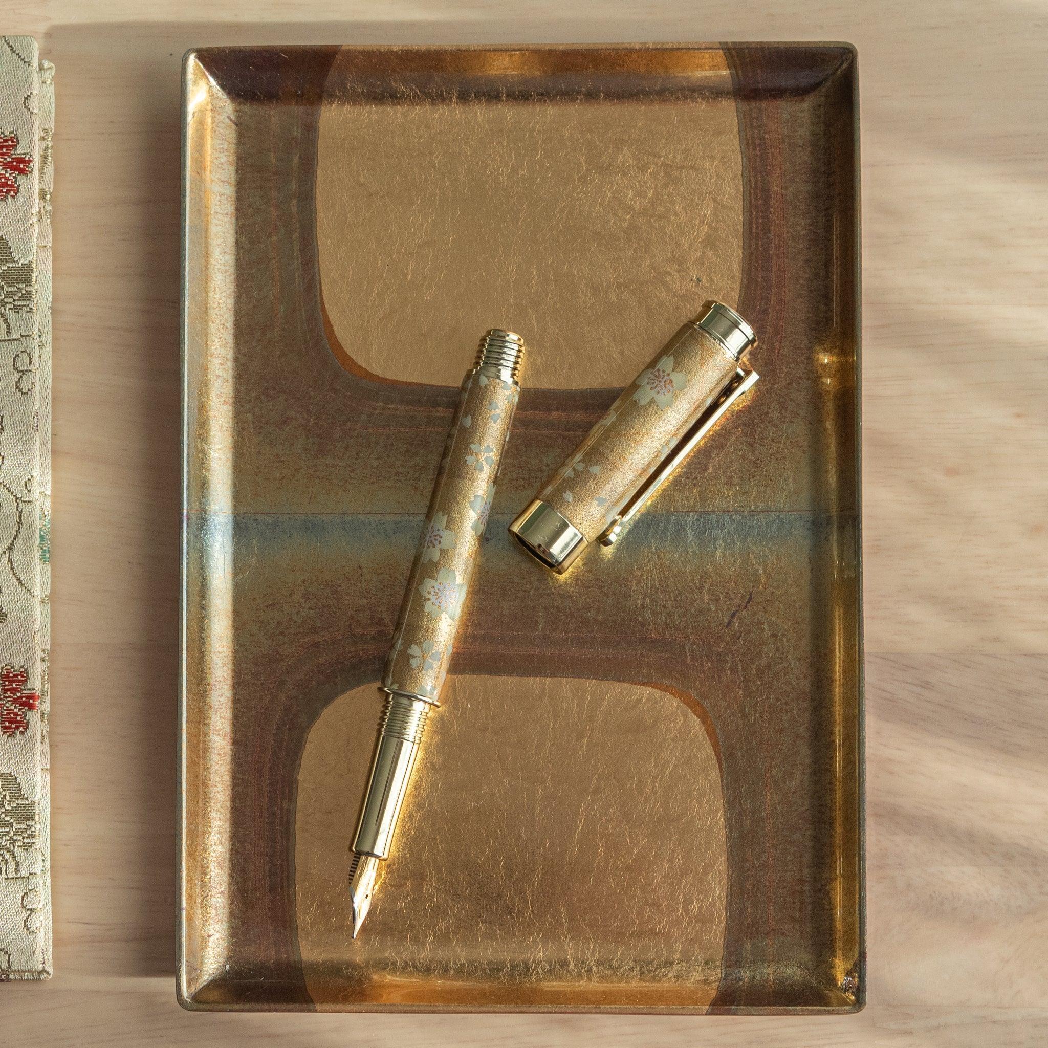 A Japanese stationery fountain pen with gold sakura details, disassembled, placed on a gold tray on a wooden surface.