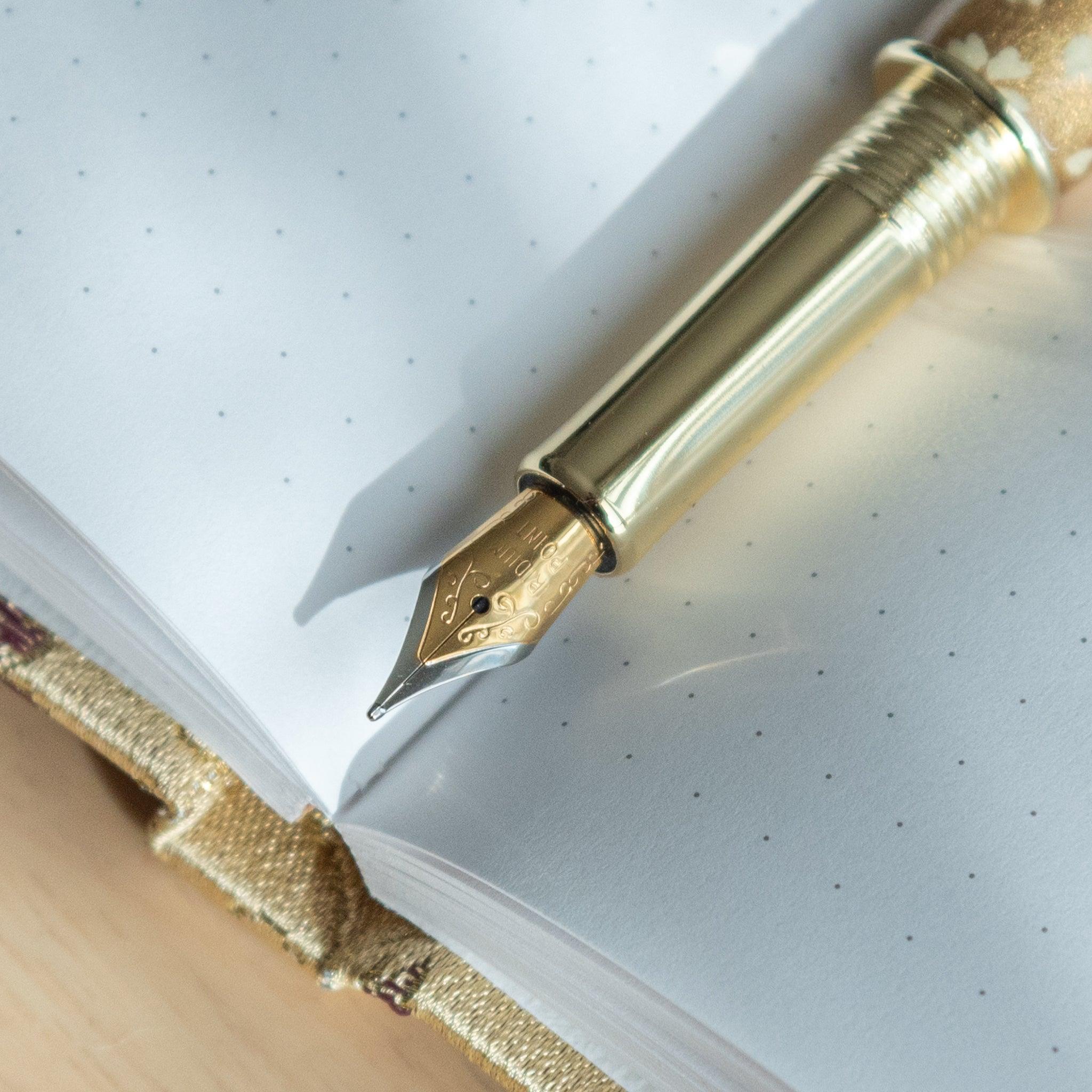 Close-up of the gold nib of a Japanese stationery fountain pen with sakura details, resting on a dotted notebook.