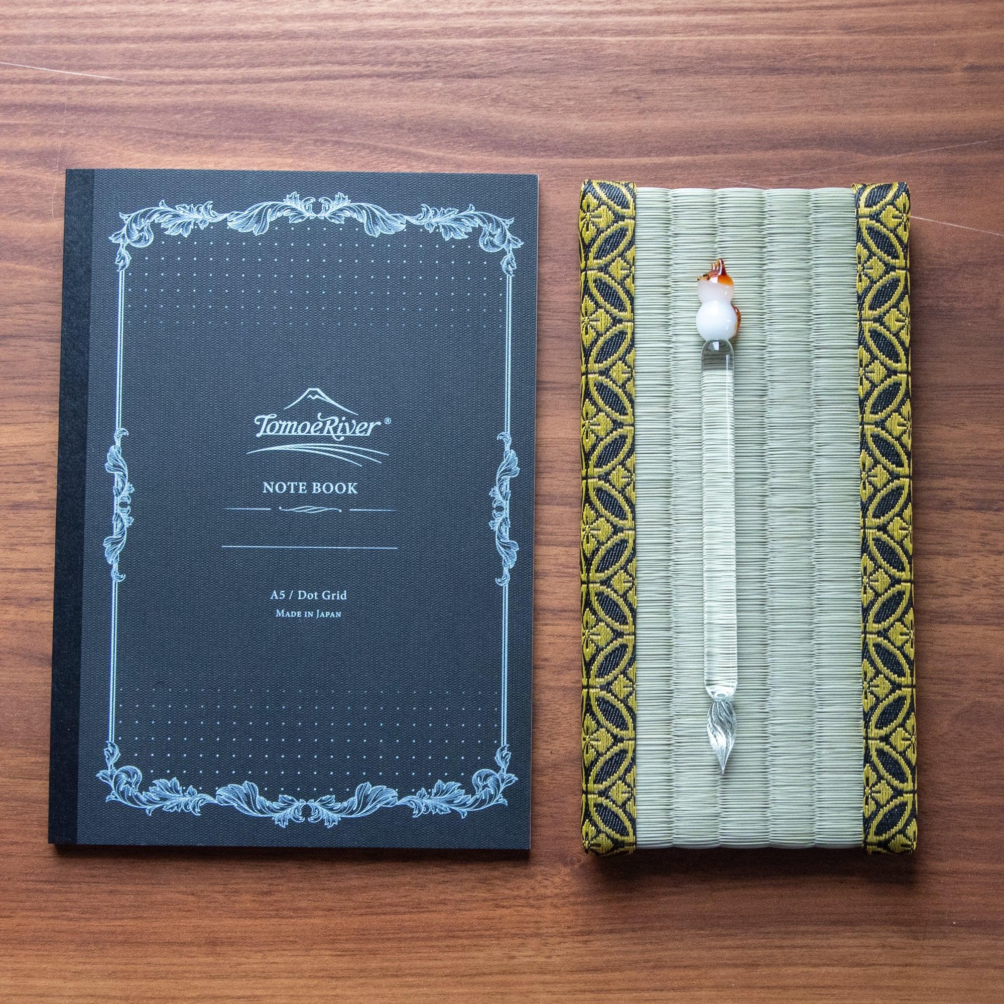 Japanese tatami pen tray with black and gold-patterned edges, showcasing its elegant woven design. Tomoe River notebook is next to the pen tray.