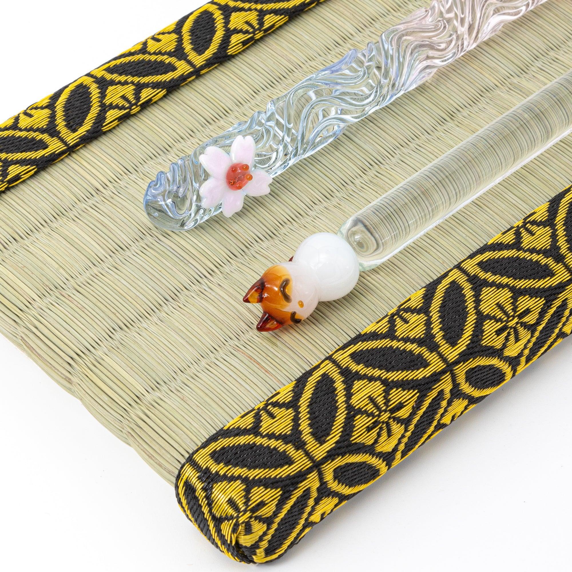 Tatami pen tray with gold-patterned borders, holding two decorative glass pens with floral and animal motifs.