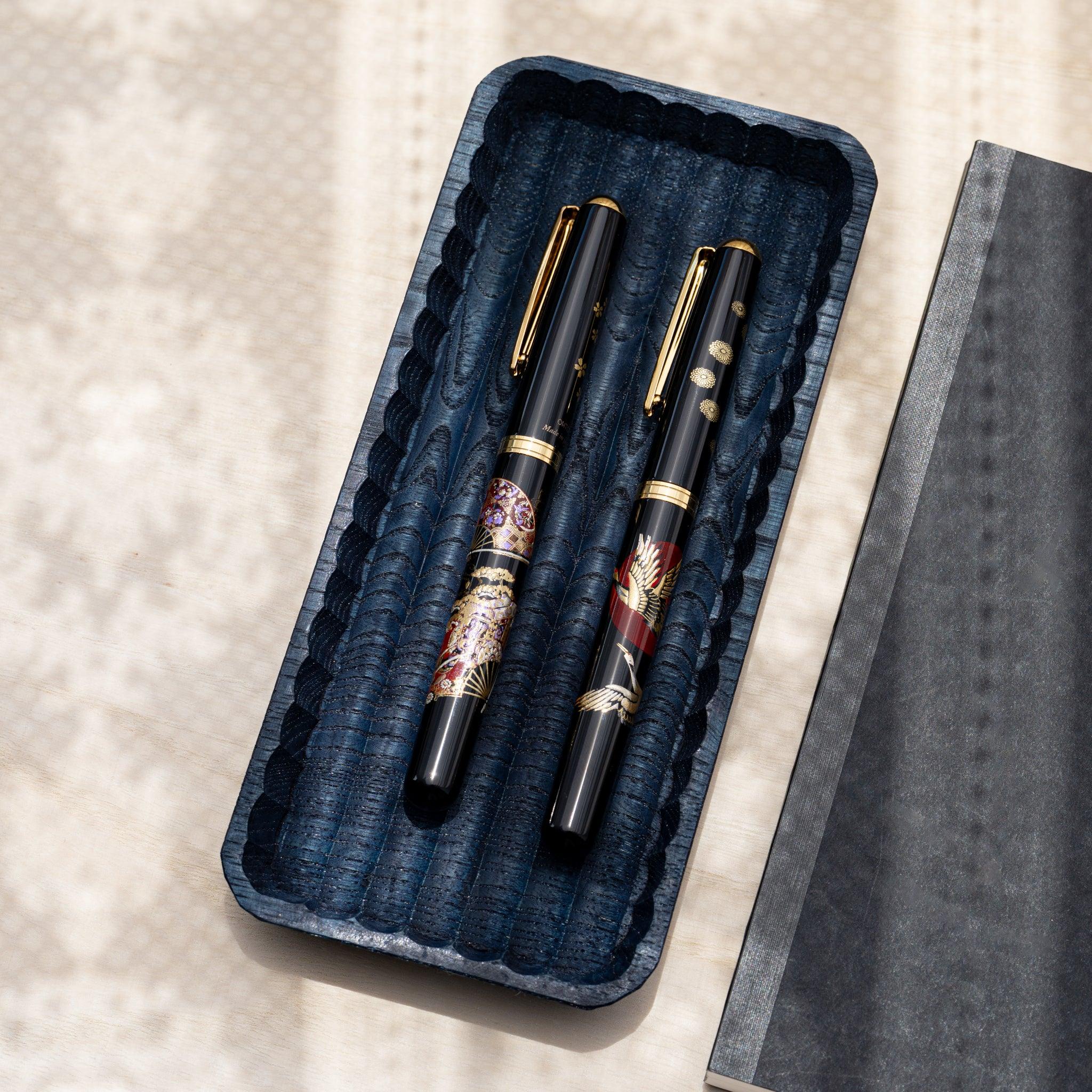 Indigo dyed slim chestnut pen tray at an angle holding two black fountain pens, dark notebook beside it