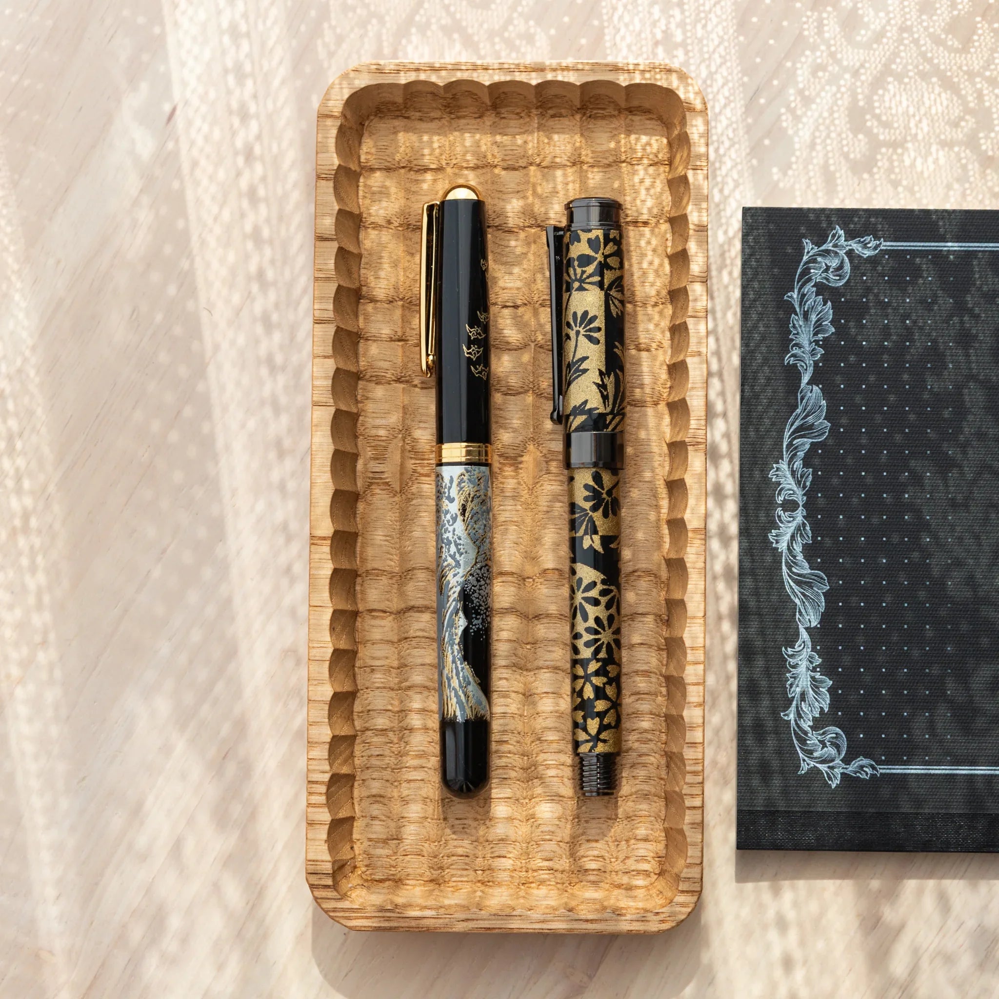 Light wood Japanese pen tray from above with parallel groove carving and two black pens in Hokusai wave and sakura designs