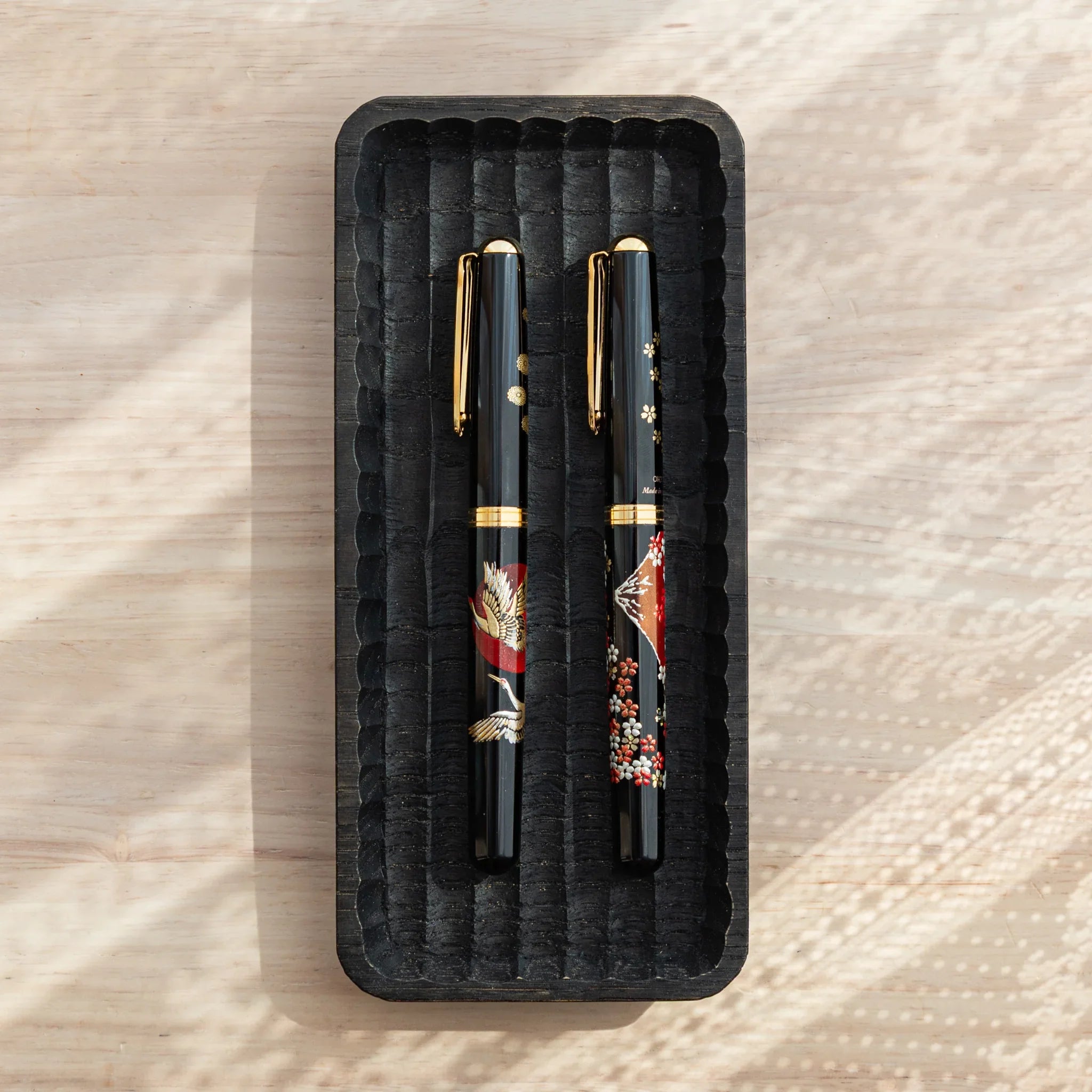 Charcoal Japanese pen tray from above with two black fountain pens in red crane and Mt. Fuji sakura designs on wood surface