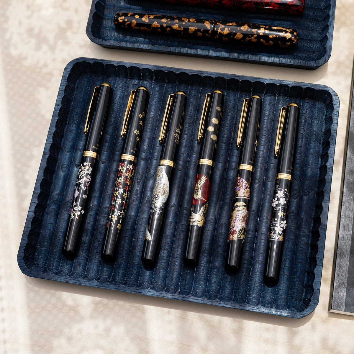 Indigo dyed wide and slim chestnut pen trays on a pale wood desk, the wide tray holding six black fountain pens
