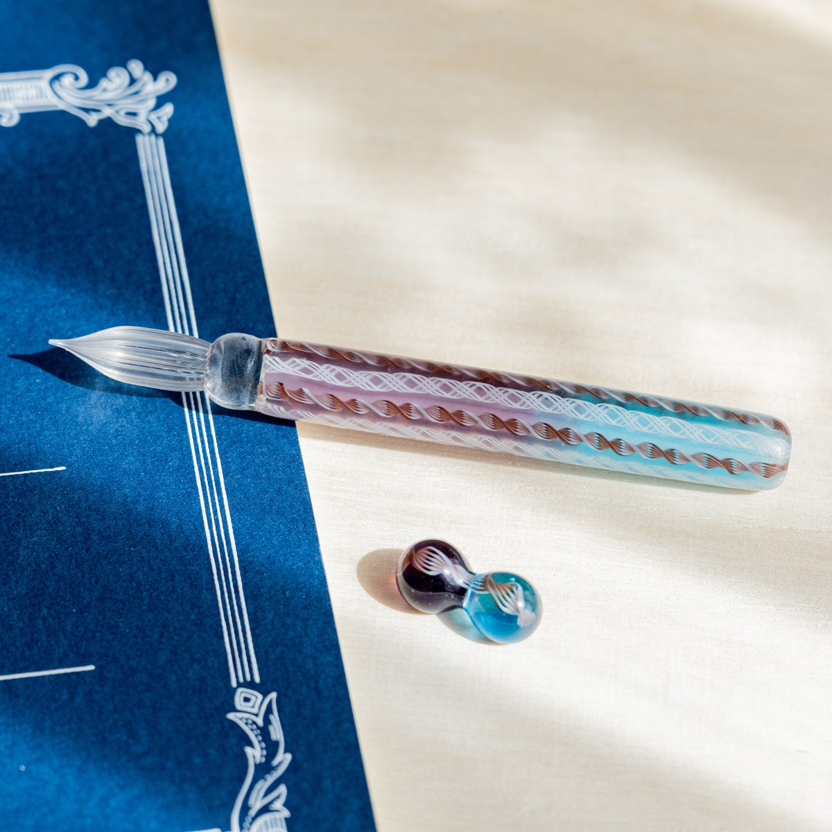 Helix Dawn glass dip pen with turquoise and dusty pink barrel beside its brown and turquoise glass pen rest