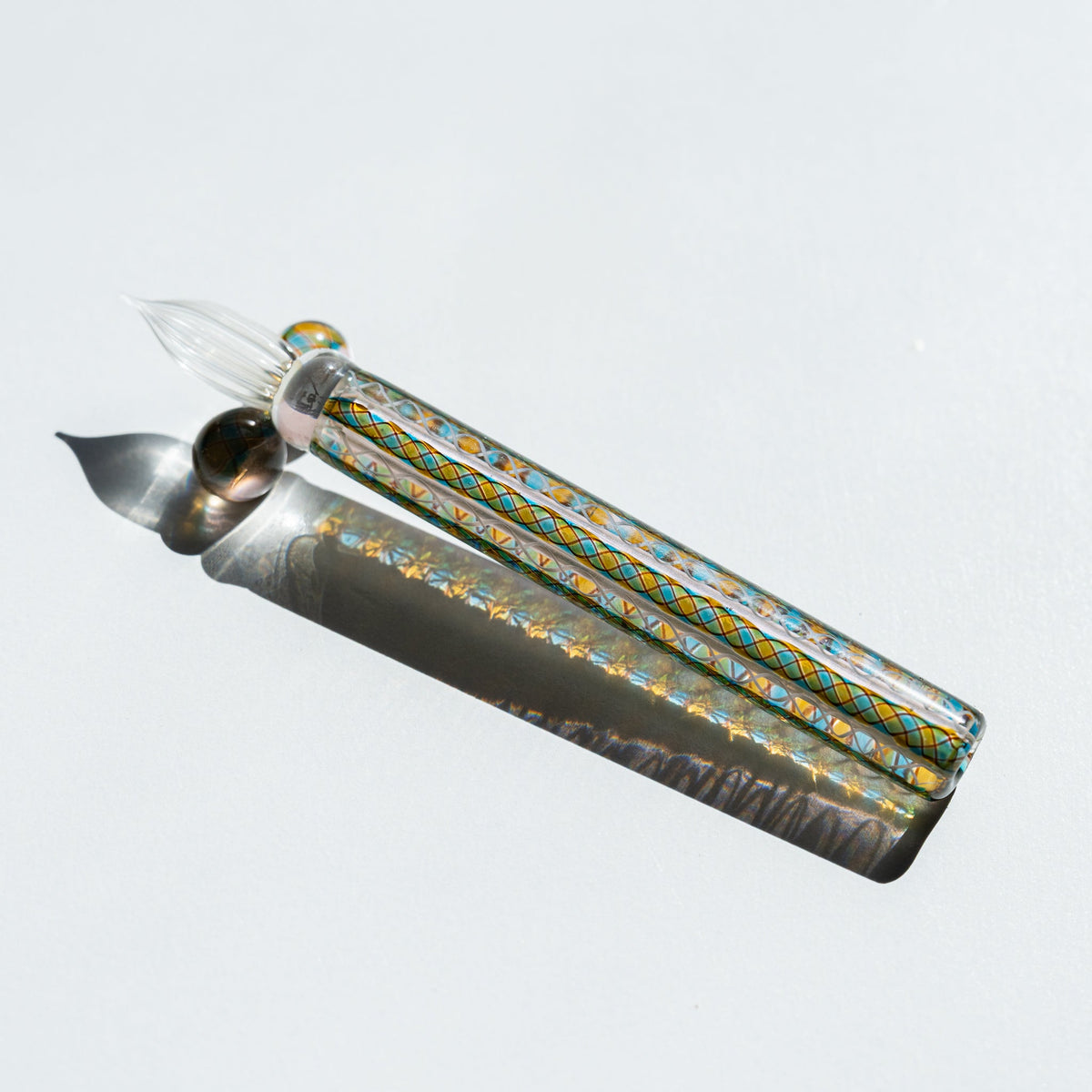 Helix Peacock glass dip pen on white, clear barrel with teal and gold lattice canes and a glass pen rest