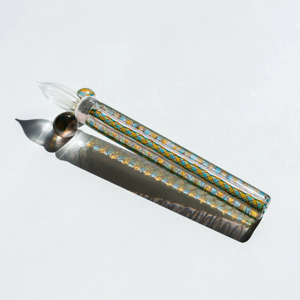 Helix Peacock glass dip pen on white, clear barrel with teal and gold lattice canes and a glass pen rest