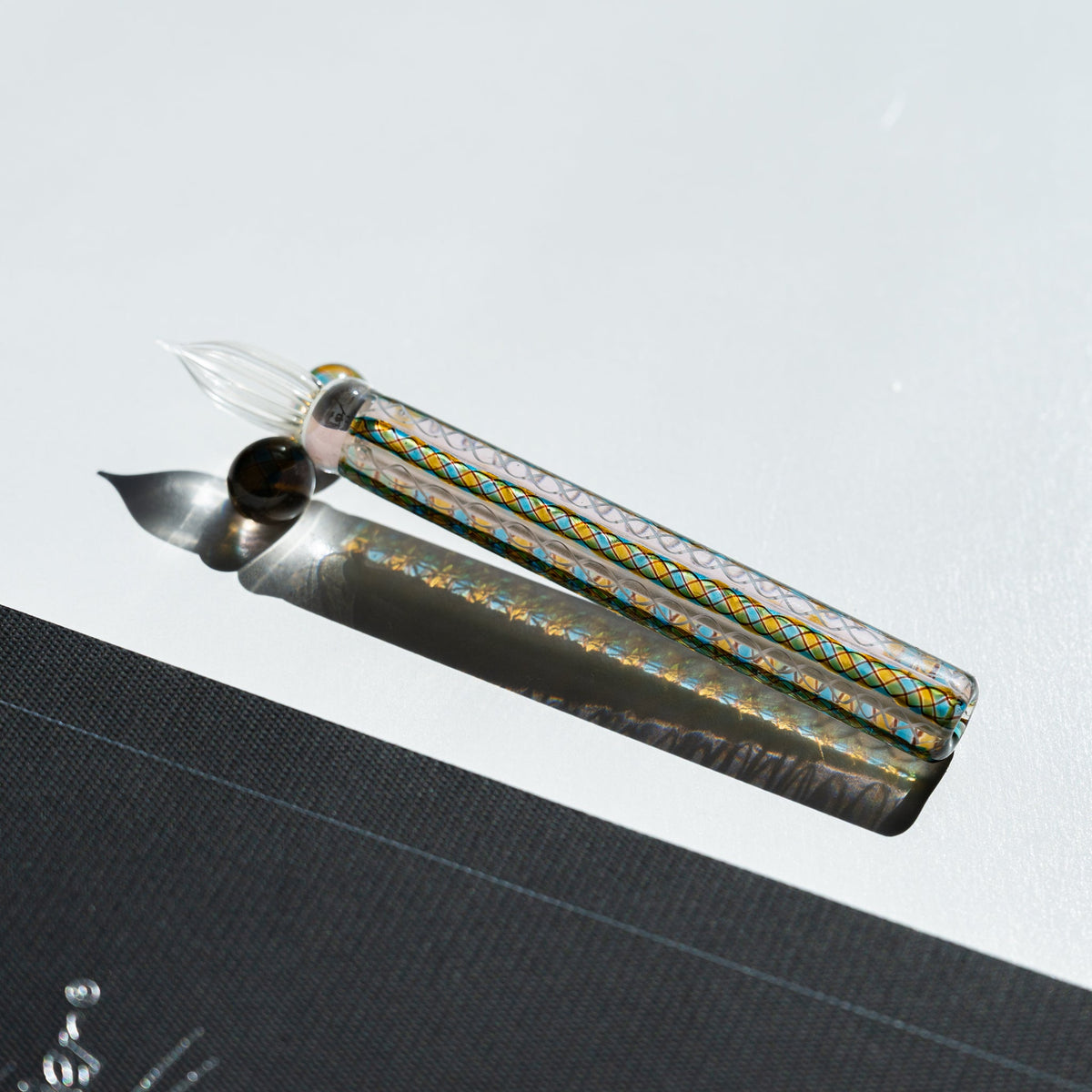 Helix Peacock glass dip pen with teal and gold cane barrel and clear fluted nib beside a dark grey card