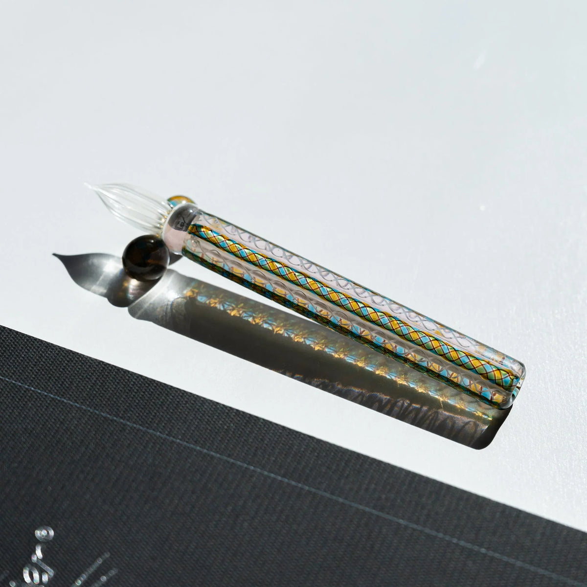 Helix Peacock glass dip pen with teal and gold cane barrel and clear fluted nib beside a dark grey card