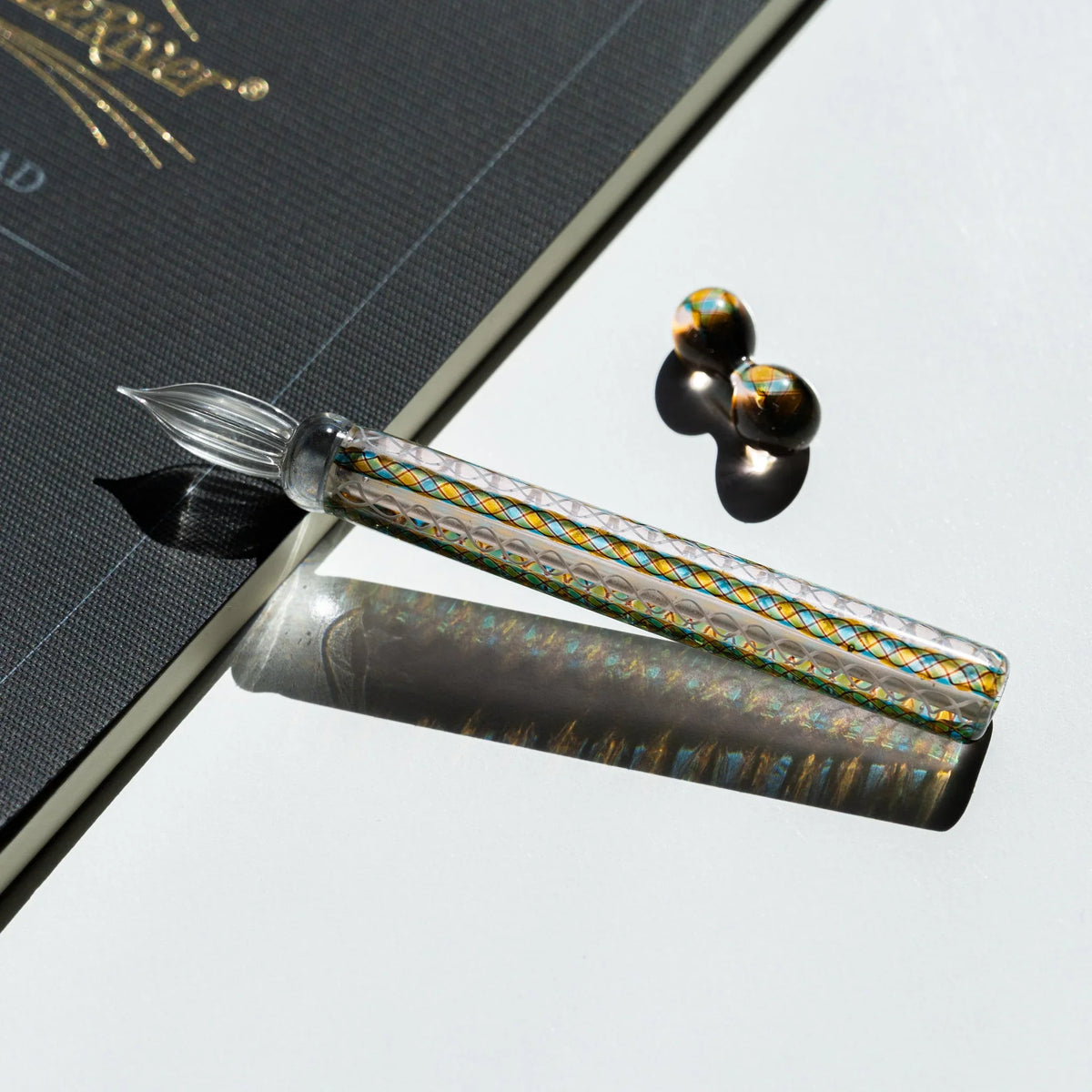Helix Peacock glass dip pen laid on the corner of a charcoal notebook with gold foil printing