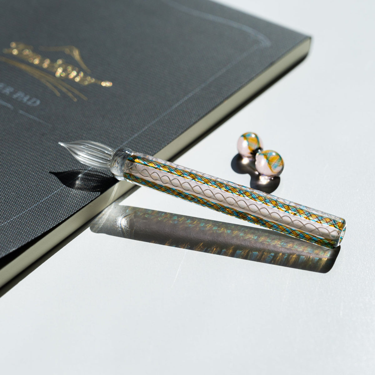 Helix Peacock glass dip pen on a charcoal notebook with its two-sphere teal and gold glass pen rest