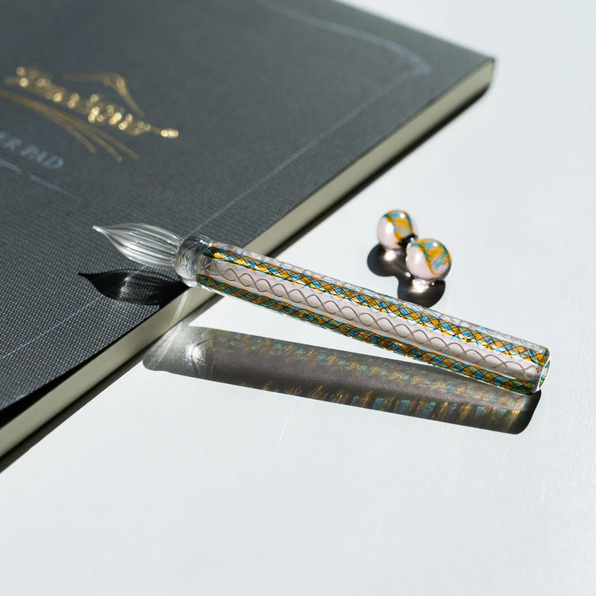 Helix Peacock glass dip pen on a charcoal notebook with its two-sphere teal and gold glass pen rest