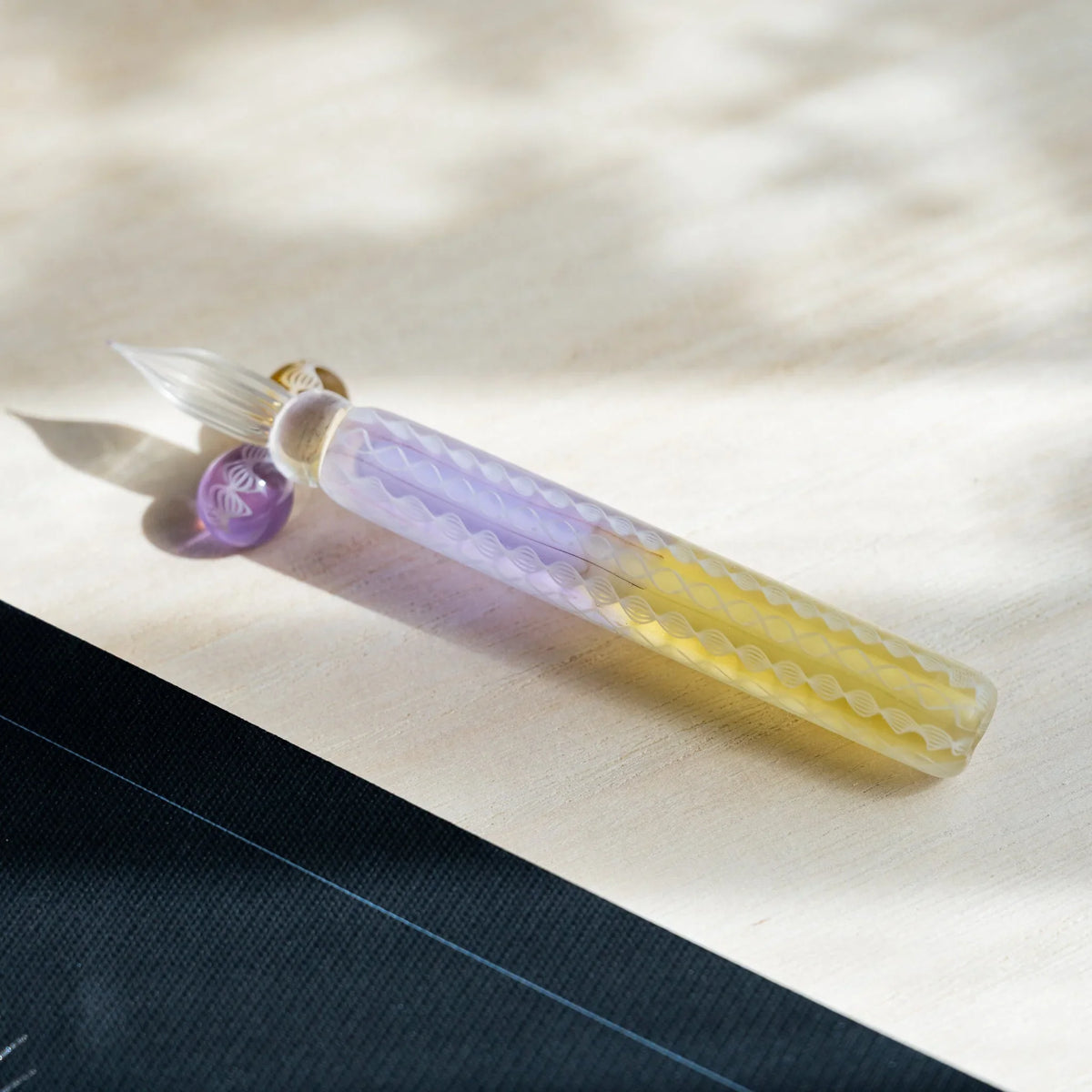 Helix Wisteria glass dip pen angled on pale wood, clear fluted nib resting on the lavender glass sphere