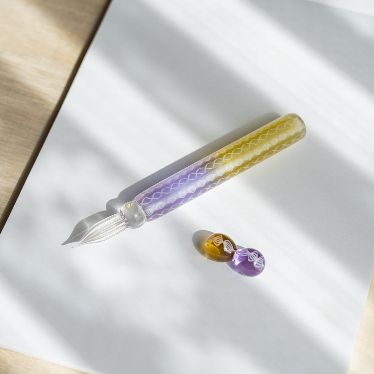 Helix Wisteria glass dip pen on white paper with its two-sphere glass pen rest in amber and lavender