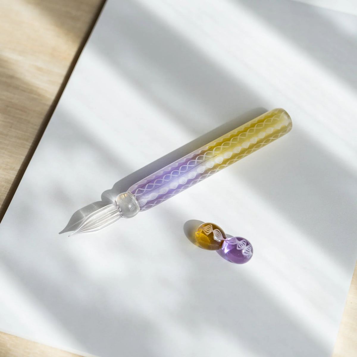 Helix Wisteria glass dip pen on white paper with its two-sphere glass pen rest in amber and lavender