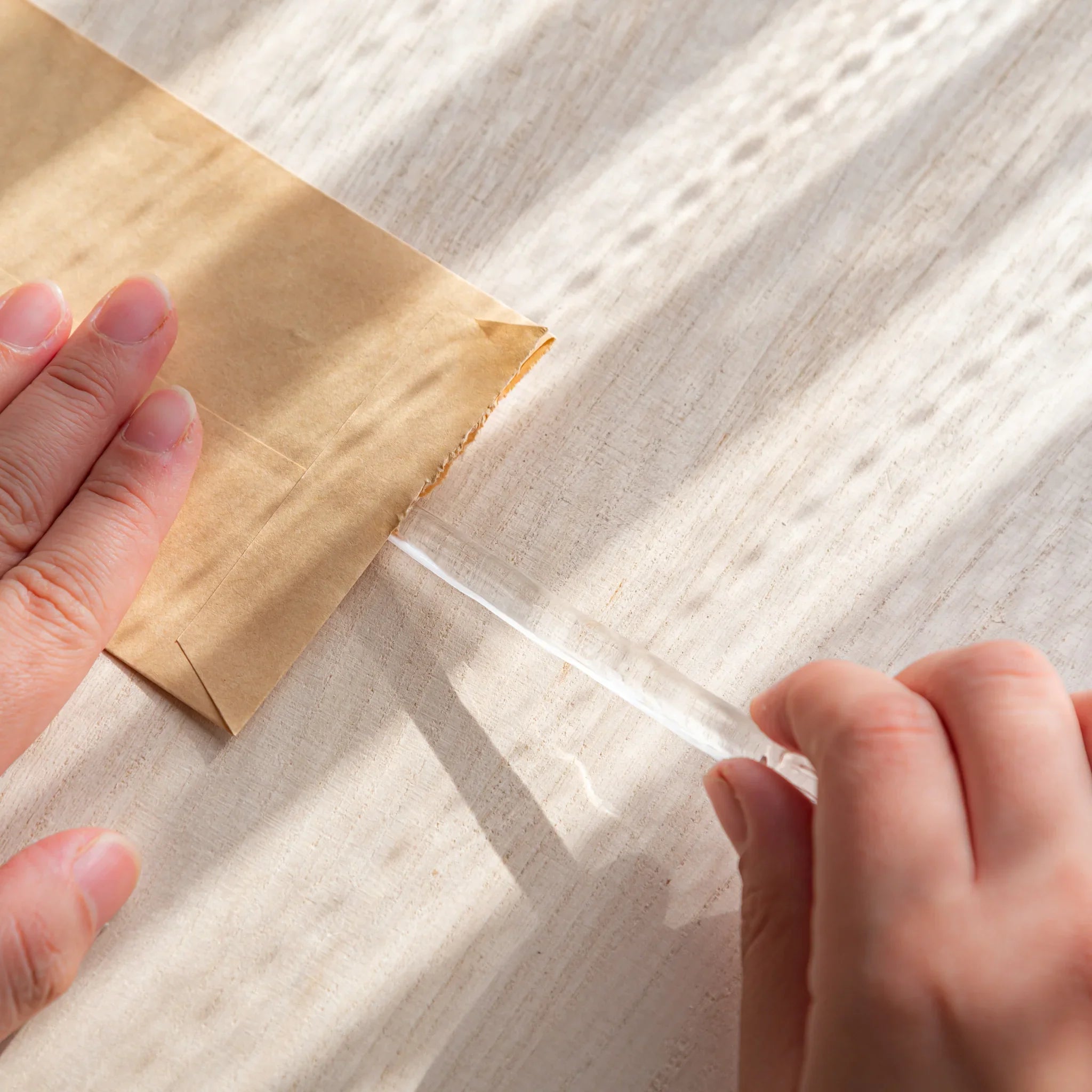 Hands opening a kraft envelope with a clear Hikari glass letter opener on a sunlit desk, featuring Japanese Stationery.