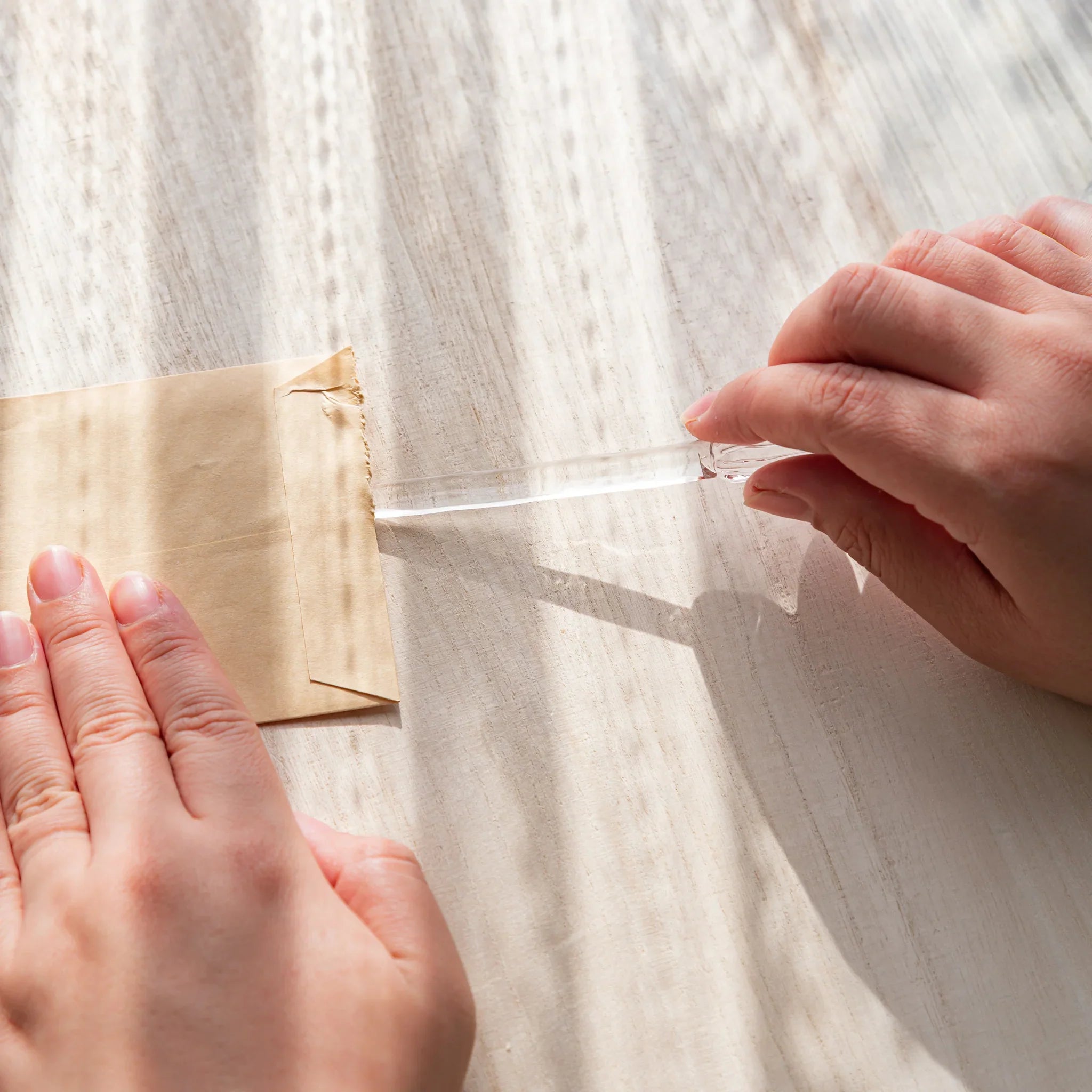 Hands opening a kraft envelope with a clear Hikari glass letter opener on a sunlit wooden desk.